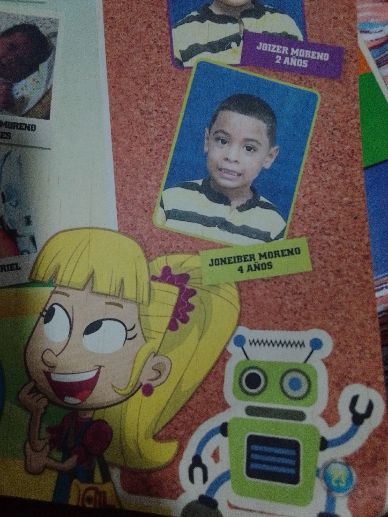The image appears to be a collage of photographs and cartoon drawings, possibly from a scrapbook or a school project. It is not possible to determine the location of La Tagua, Venezuela, from the provided image.
The top portion of the image features a photograph of a young child, likely a toddler, wearing a bib. To the right of this photograph, there is a cartoon drawing of a child with a striped shirt and blonde hair, along with a purple label that reads "JOIZER MORENO 2 AÑOS."
Below this, there is a photograph of a young boy with short, dark hair and a striped shirt. A yellow label beneath him reads "JONEIBER MORENO 4 AÑOS."
The lower left of the image shows a cartoon drawing of a girl with blonde hair, large eyes, and a wide smile. She is wearing a red shirt and blue overalls and appears to be holding a yellow bag.
To the right of the girl, there is a cartoon drawing of a green robot with blue accents. The robot has two circular eyes, a rectangular body with a display, and articulated arms and legs.
The background of the collage is a textured, reddish-brown surface. The overall presentation suggests a collection of personal mementos related to children.