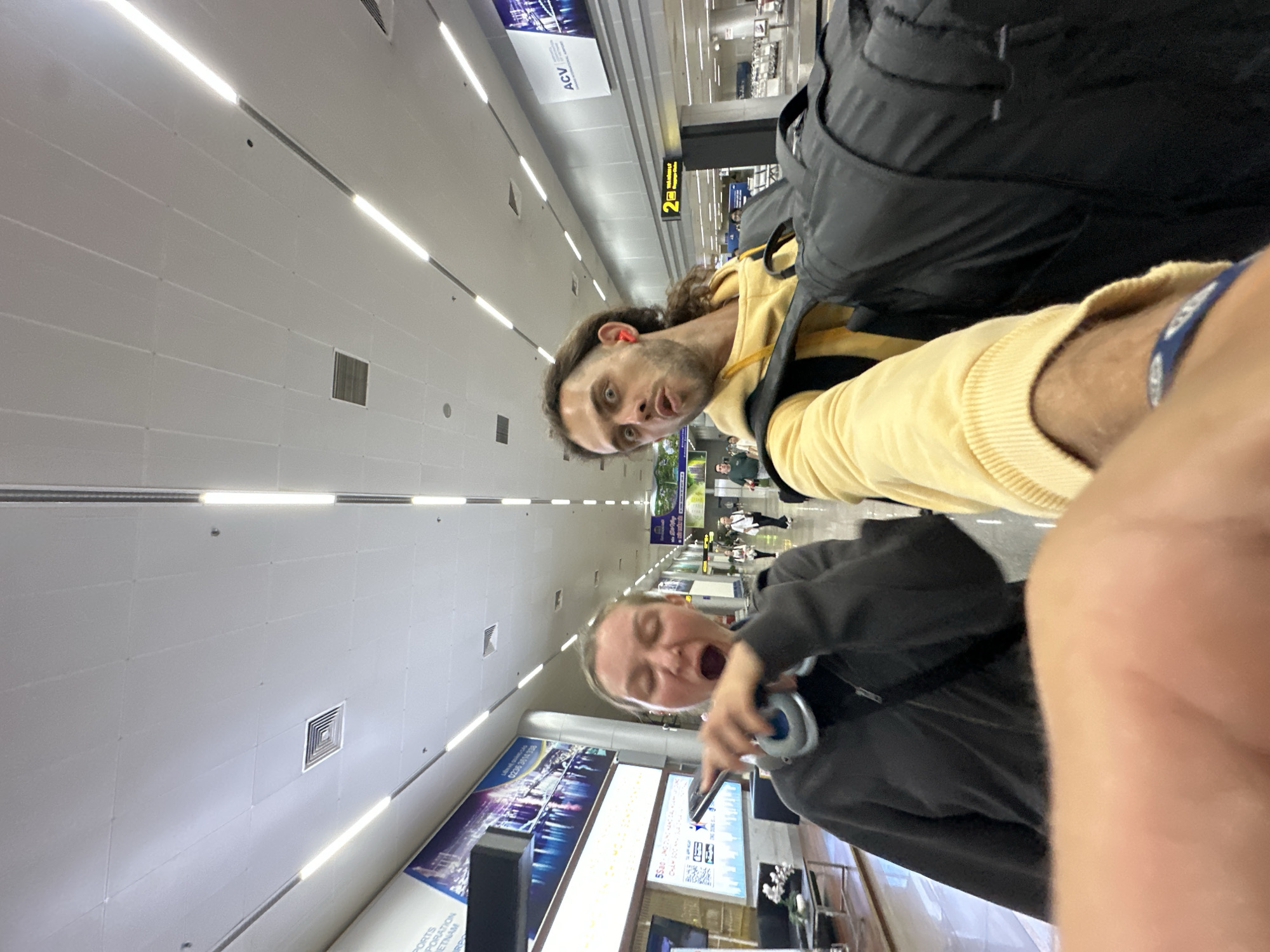 The image depicts a selfie taken by two individuals in an airport. The person on the left, a woman, is wearing a dark jacket and has large headphones around her neck. Her mouth is open in what appears to be a yawn or a loud exclamation, and she is holding a phone in her right hand, pointing it outwards. The person on the right, a man, is wearing a yellow hoodie and has a black backpack slung over his shoulder. He is making a surprised or playful facial expression.
The background reveals the interior of an airport terminal. The ceiling is white with fluorescent lighting. There are large advertisements on the walls. Signage in Vietnamese is visible, indicating the location is an airport in Đà Nẵng, Vietnam. One sign reads "2" followed by Vietnamese text related to baggage claim. Other passengers can be seen in the distance, as well as baggage carousel equipment. The overall scene suggests the individuals are at the airport, possibly waiting for a flight or collecting luggage.