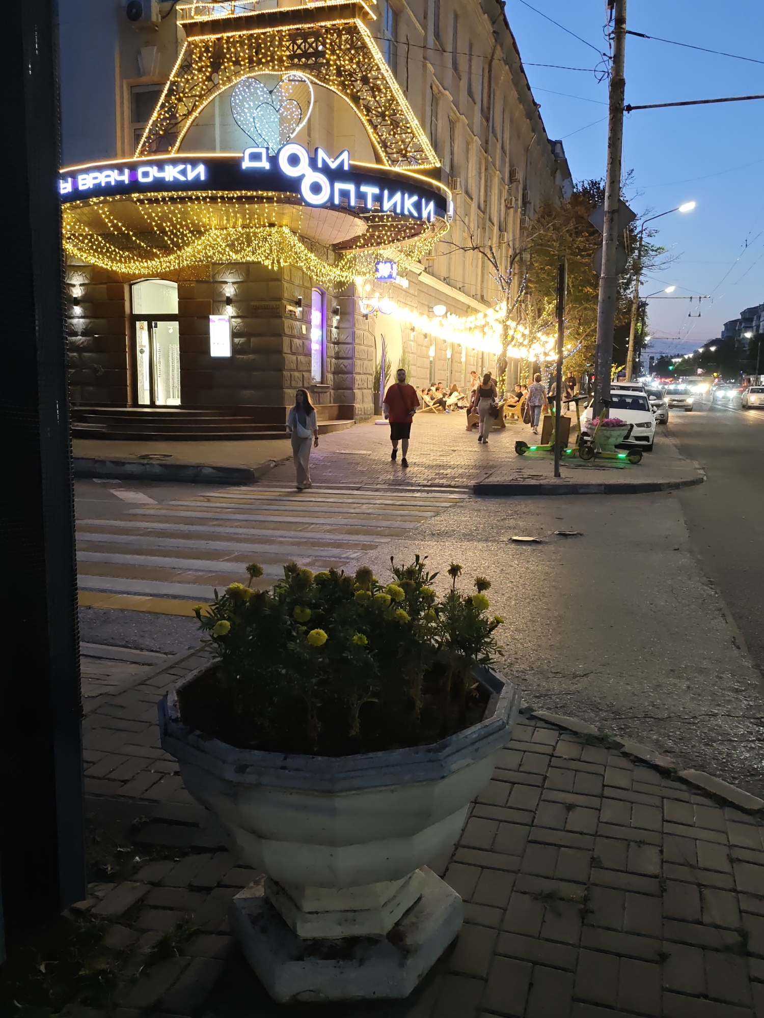The image depicts a street scene in Rostov-on-Don, Russia, at dusk. A building with elaborate golden lights outlining its facade and a large heart motif is prominently featured. The building appears to be a commercial establishment, with signage in Cyrillic that reads "ДОМ ОПТИКИ" (Dom Optiki, meaning Optical House) and "ВРАЧ ОЧКИ" (Vrach Ochki, meaning Eye Doctor).
A pedestrian crossing is visible in the foreground, with a woman in light-colored clothing walking across it. To the right of the street, there are several parked cars, two electric scooters with illuminated accents, and a line of people seated at outdoor tables, likely at a cafe or restaurant. A large concrete planter filled with yellow flowers is situated on the sidewalk in the foreground. The sky is a deep blue, indicating twilight. The street is paved, and there are streetlights illuminating the area. The overall impression is that of a lively urban environment as evening sets in.