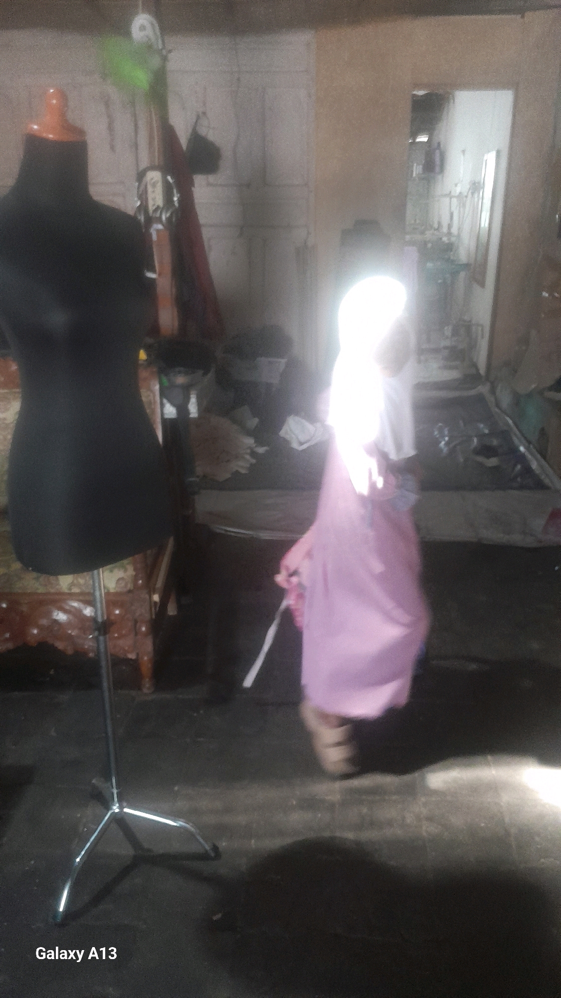 In a dimly lit indoor space, possibly a workshop or storage area, a black fabric mannequin stands to the left of the frame. The mannequin is positioned on a metal stand with three legs. To the right, a person is visible, wearing a light-colored hijab and a pink dress. They appear to be in motion, possibly walking or posing. The background is cluttered with various objects, including what looks like clothing racks, fabric, and possibly furniture, suggesting a place where sewing or garment production might occur. A doorway or opening on the right side of the image leads to another area with what appear to be shelves and more items. The floor is dark and appears to be concrete or a similar material. The overall impression is of a functional, somewhat unorganized space. The metadata indicates the image was taken in Ngelo, Indonesia.