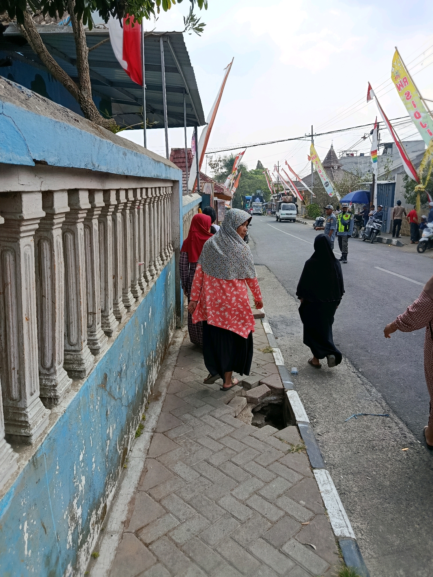 A street scene in Gubukklakah, Indonesia, with a bright blue wall on the left side of the image. The wall has a decorative white balustrade running along its top. Several people are walking on a paved sidewalk next to the wall. One person in the foreground is wearing a red and white floral pattern top and a black skirt, with a patterned headscarf. Behind them, another person in a black headscarf and black dress is walking on the sidewalk, and a third person in a red headscarf and plaid shirt is also visible. Further down the street, there are more people, including a person in a yellow safety vest. Various flags and banners are flying along the street, some with red and white colors, suggesting a festive or national event. The street is paved, and there are vehicles including cars and motorcycles. The sky is overcast. A section of the sidewalk to the left of the person in the black dress has collapsed, creating a hole.