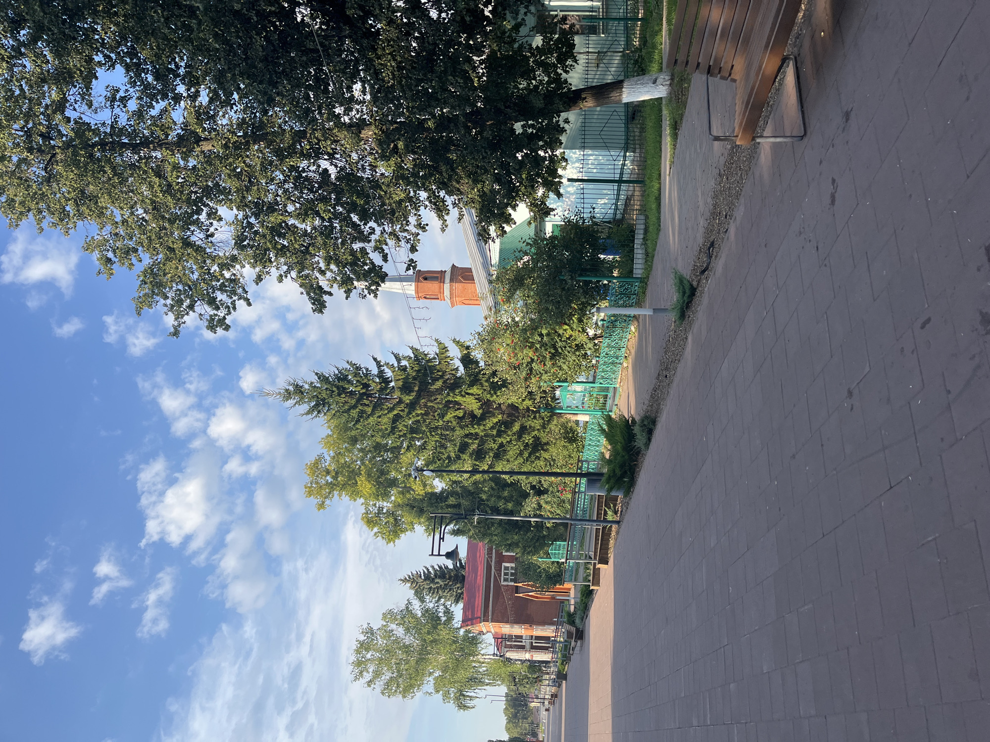 The image shows a paved walkway in a town square, with trees and buildings on the right side. The walkway is made of dark gray rectangular pavers. On the right side of the walkway, there is a wooden bench with metal legs. Behind the bench is a green metal fence and a building with light blue walls and a green roof. Further back, there is a brick building with a red roof and a tall brick tower with a spire. The sky is blue with scattered white clouds. The overall scene is serene and depicts a public space in the town of Novye Turbasly, Russia, likely on a clear day.