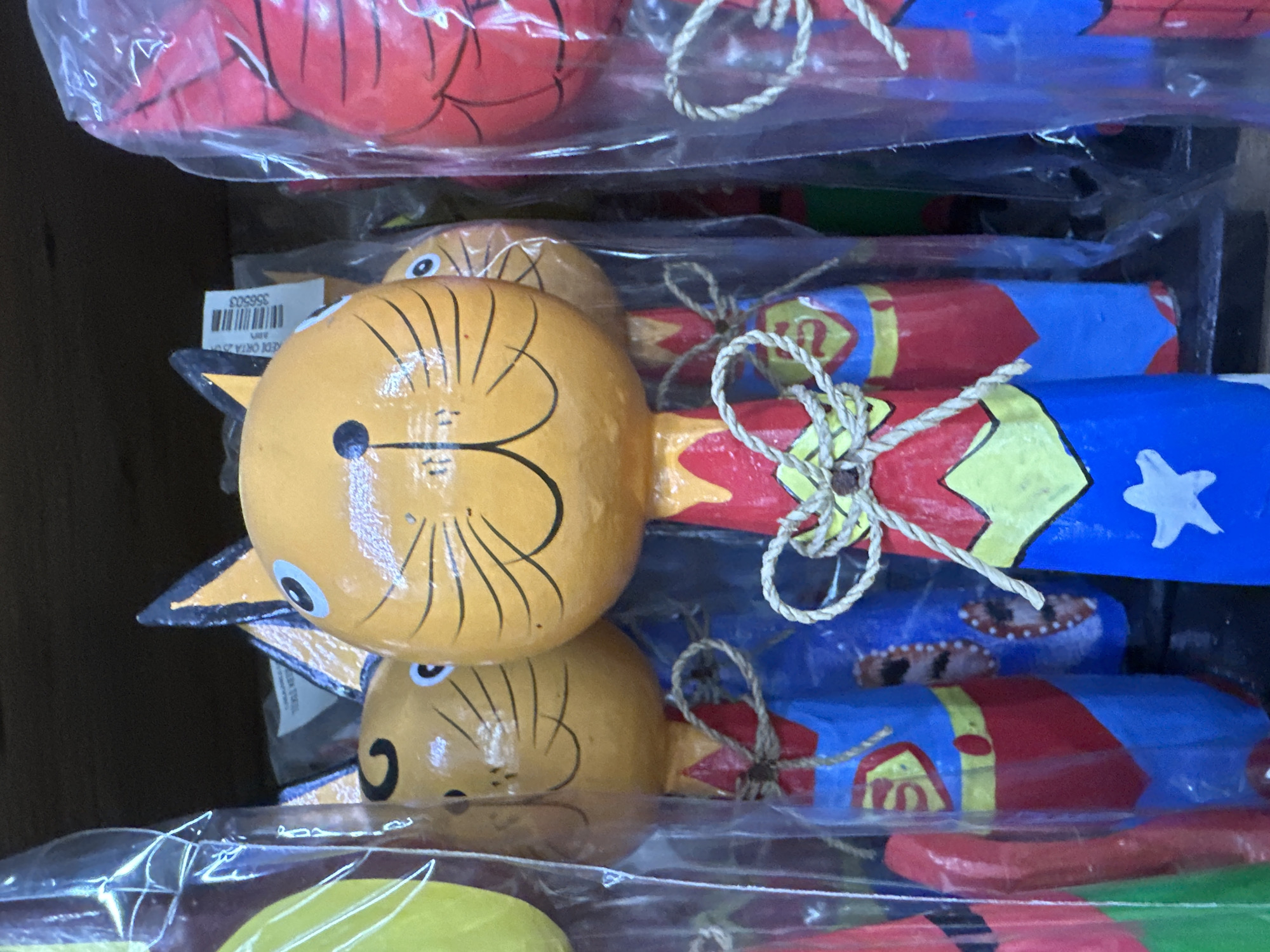 The image shows a stack of packaged, handmade wooden toy fish. The fish have round, yellow heads with black facial features resembling cats. Their bodies are elongated and painted with colorful designs, including blue, red, and white star patterns. Each fish is tied with a piece of twine. The toys are enclosed in clear plastic packaging. A barcode sticker is visible on one of the packages. The scene is most likely in a shop or market in Fethiye, Turkey, given the style of the toys.