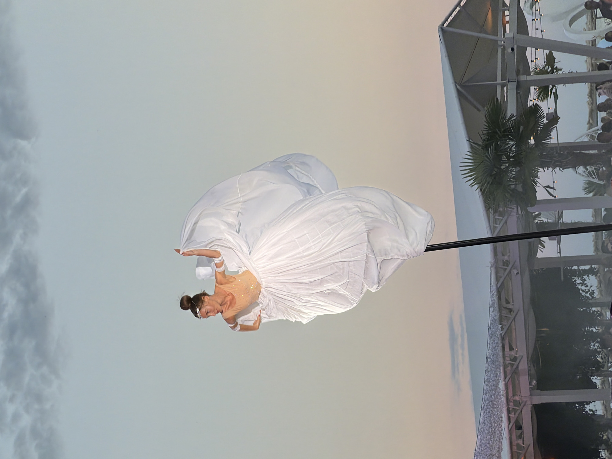 A performer in a white dress with a sequined bodice stands on a pole, elevated against a pale sky at sunset. The dress billows dramatically around her, resembling a large flower or cloud. She wears a jeweled headband. Below her, a white tent structure with a thatched roof is visible, along with palm trees and a crowd of people. The scene appears to be at an outdoor venue, possibly a beach or resort, in Odesa, Ukraine, given the context provided. The sky shows soft hues of pink and orange from the setting sun, with scattered clouds.