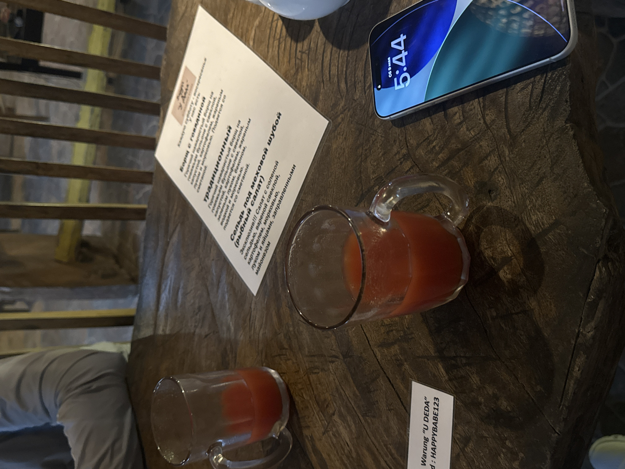 Here is a description of the image:
**Location:** The image was most likely taken at a restaurant named "U DEDA", in the city of Kuta Selatan, Indonesia. This is inferred from the text on a small card in the bottom right corner of the image which reads "Warung 'U DEDA' - d: HAPPYBABE123." Warung is an Indonesian word meaning "small shop" or "stall", and is often used to refer to restaurants.
**Scene:** The image shows a wooden table with several items on it. There is a menu, a smartphone displaying the time as 5:44, a glass mug containing an orange liquid, and another glass mug with a smaller amount of the same liquid. The light suggests it might be a dining table, near a person sitting and/or dining. The picture appears to be taken from an elevated perspective.
**Objects:**
* Menu with text in Russian.
* Smartphone with a blue and white screen.
* Glass mugs filled with an orange liquid, possibly juice or a mixed drink.
* Wooden table with a visible wood grain pattern.
* A person's leg and a part of a chair.
**People:**
* Only a part of a person's leg is visible. No other individuals are identifiable.
**Actions:**
* No explicit actions are depicted in the image. It appears to be a static scene.
**Interactions:**
* There are no apparent interactions between individuals or objects within the frame.
**Environment:**
* The setting seems to be a restaurant or cafe.
* The lighting appears natural, suggesting it is daytime.