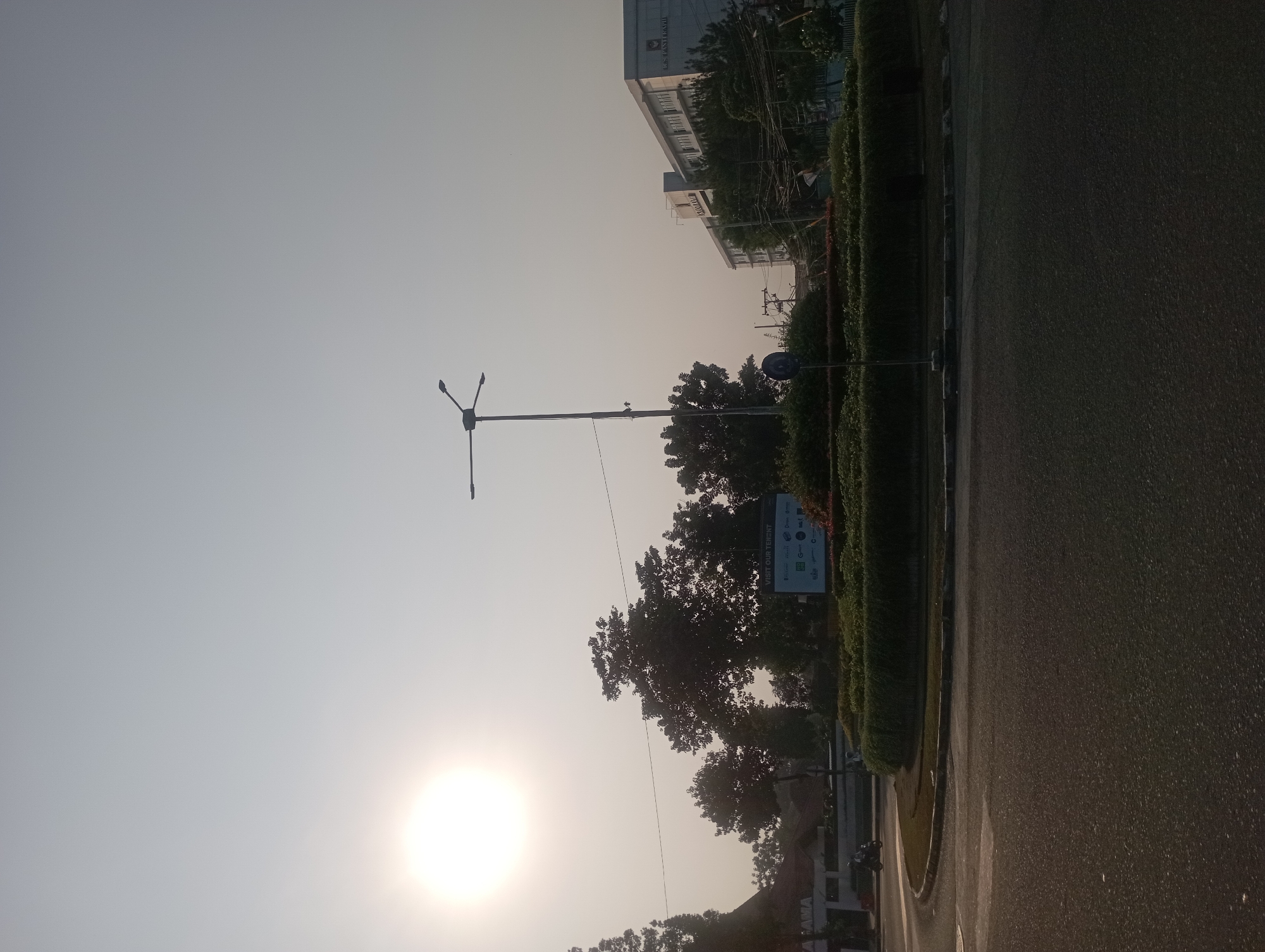 The image captures a bright, hazy day in Depok, Indonesia, likely during the early morning or late afternoon, given the low angle of the sun. The sun, a large, luminous disc, dominates the upper left quadrant of the frame, casting a soft glow that contributes to the overall muted color palette.
In the foreground, a paved road curves gently, suggesting a roundabout or intersection. The road surface is asphalt, with some visible texture and slight shadowing.
To the right, a circular green space, likely a traffic island or park median, is densely planted with low-lying shrubs and bordered by a decorative red flowering plant. A modern streetlamp with three arms stands tall in the center of this green area.
Behind the green space, a multi-story building, possibly a commercial establishment or office, stands prominently. It is light-colored with visible windows and signage. The name "RS PANTI RAH..." is partially legible on its facade, indicating it might be a hospital. Further to the left, another building with a reddish-brown roof and partial signage reading "AMA" is visible.
Several trees with dark green foliage are scattered throughout the scene, some appearing mature and full. Power lines are strung between poles, contributing to the urban landscape.
In the middle ground, a white billboard with multiple logos is placed behind the green circular planting. A sign that reads "VISIT OUR TENANT" is visible at the top of the billboard.
A lone motorcyclist is seen on the road to the left, adding a sense of movement to the otherwise static scene. The sky is a pale, almost white haze, indicating atmospheric conditions that obscure sharper details and contribute to the diffused lighting. The overall impression is one of a busy urban environment during a time of soft, diffused light.