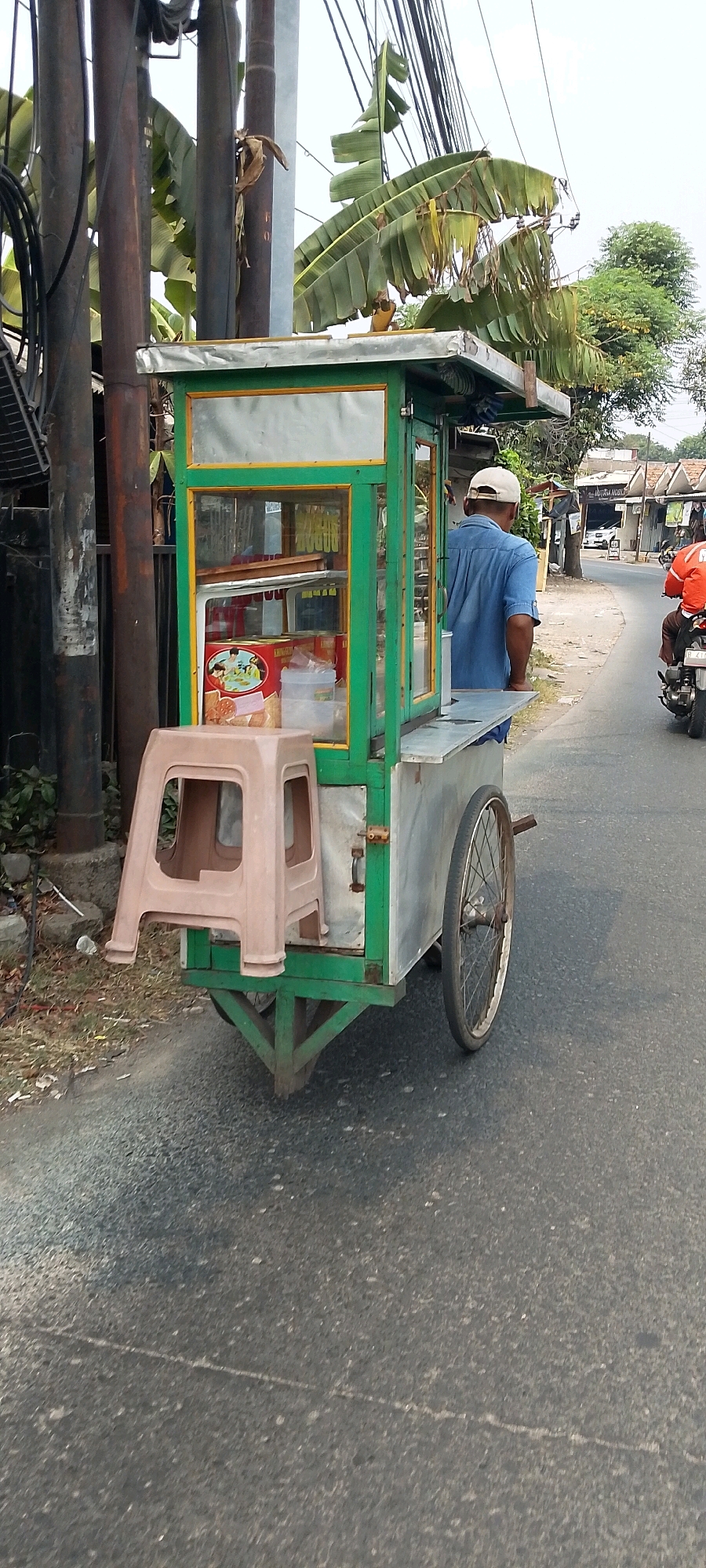 In a street scene in Bekasi, Indonesia, a vendor operates a mobile food cart. The cart, painted green and yellow, is equipped with glass display cases showing various packaged goods, including boxes of crackers and other snacks. A beige plastic stool is placed on the front of the cart. A man wearing a blue shirt and a white cap stands behind the cart, appearing to be the vendor. The cart has large spoked wheels and is situated on an asphalt road. In the background, a person on a motorcycle with an orange jacket is visible further down the road, and several buildings line the street. Banana trees with large leaves are prominent on the left side of the image, alongside utility poles and electrical wires, suggesting a semi-urban or suburban setting. The overall scene depicts a typical street-side vending operation.