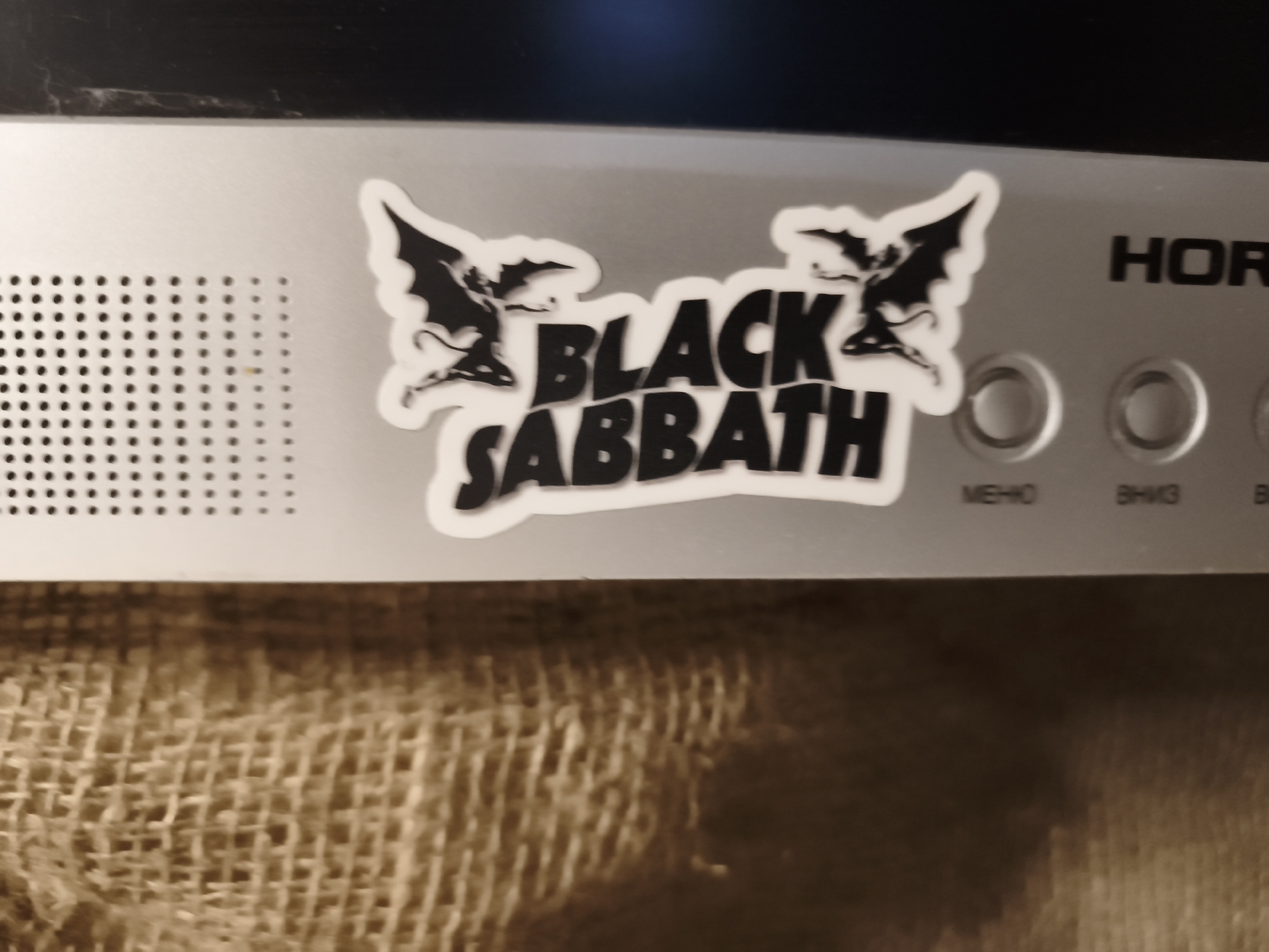 The image displays a close-up, low-angle shot of a silver-colored electronic device, possibly a guitar amplifier or audio equipment, with a Black Sabbath band sticker prominently placed on its surface. The sticker features the band's logo in a stylized font, accompanied by two winged demonic figures on either side. The device has a perforated section on the left and several circular controls with Cyrillic labels ("МЕНЮ", "Вниз", etc.) on the right. The background is a textured, woven material, likely burlap or a similar fabric, which forms the base of what appears to be the lower part of a speaker cabinet. The lighting suggests an indoor setting, and the overall image quality is somewhat grainy. The location is identified as Гаранскі сельскі Савет, Belarus. No people are visible in the image.