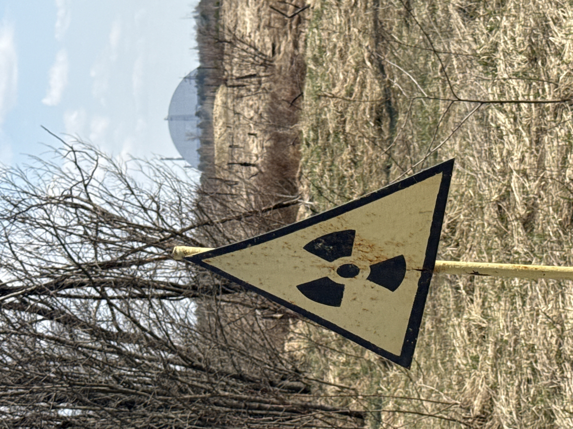 The image shows a radioactive warning sign in Хутор, Ukraine. The sign is triangular, with a yellow background and a black border. The radiation symbol is in the center of the sign. The sign is attached to a metal pole. In the background, there is a barren landscape with dead trees, vegetation, and a large structure. The sky is visible in the upper left corner of the image.