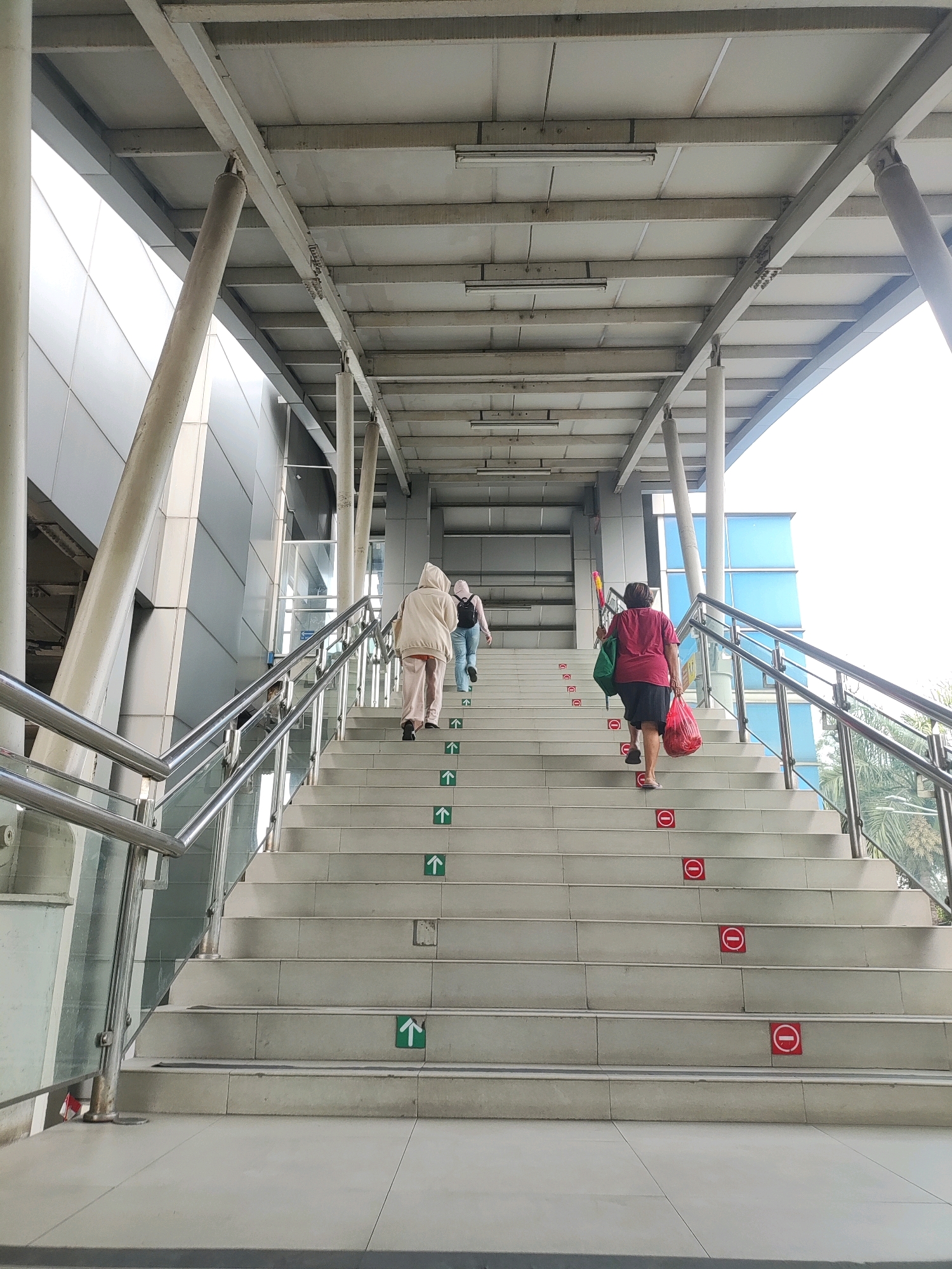 An overhead view captures a pedestrian bridge with a wide staircase. Three individuals are ascending the stairs. The person in the foreground, wearing a red shirt and dark skirt, carries two plastic bags, one red and one green. Behind them, a person in a light brown hooded outfit walks up, followed by another person with a backpack. Green arrow stickers indicating upward movement are visible on some steps, interspersed with red circular stickers featuring a diagonal line, suggesting a prohibition. The staircase is bordered by metal railings and glass panels. The surrounding environment includes modern architecture with gray metallic siding and blue glass windows. The scene appears to be located at the pedestrian overpass of the Jakarta Garden City Mall in East Jakarta, Indonesia.