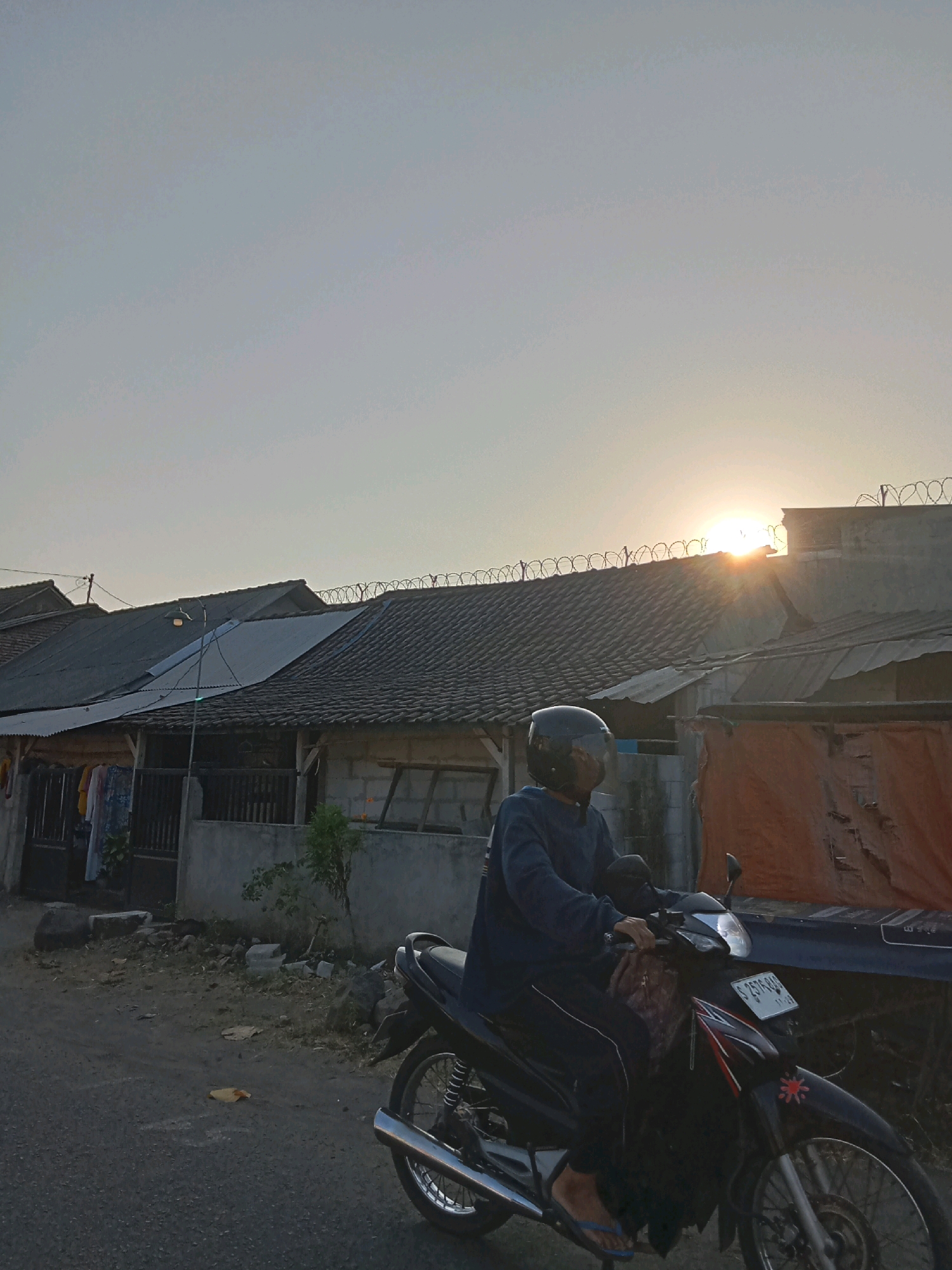 In Jombang, Indonesia, the sun sets behind houses with tiled roofs. A person wearing a blue sweatshirt and helmet rides a black motorcycle on a paved road. The motorcycle has a license plate that reads "S 2570 RAA". Houses in the background have barbed wire on their roofs for security. Clothes are hanging on a line outside one of the houses. The scene is captured during the golden hour, with the sun casting a warm glow.