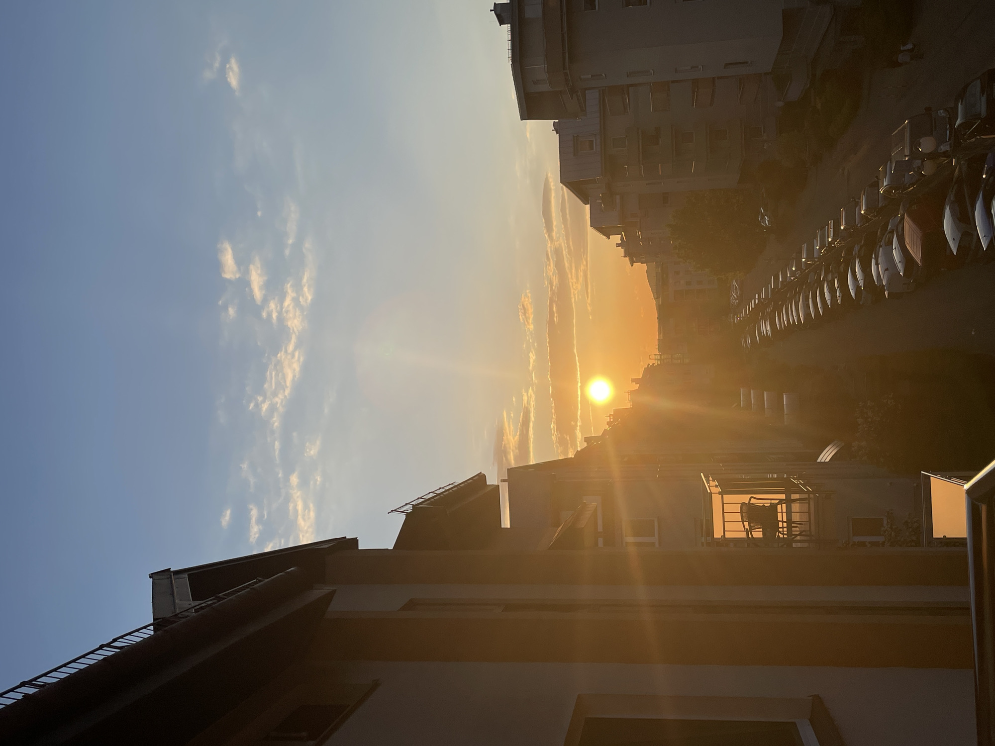 The image captures a sunset in an urban setting. The sun is positioned in the center of the frame, casting a golden light across the scene. Buildings are visible on both sides, with a line of parked cars on one side. The sky shows a gradient of blue to orange with scattered clouds. A balcony with a chair is visible in the lower right. The location is in a city, but the specific city is unknown.
