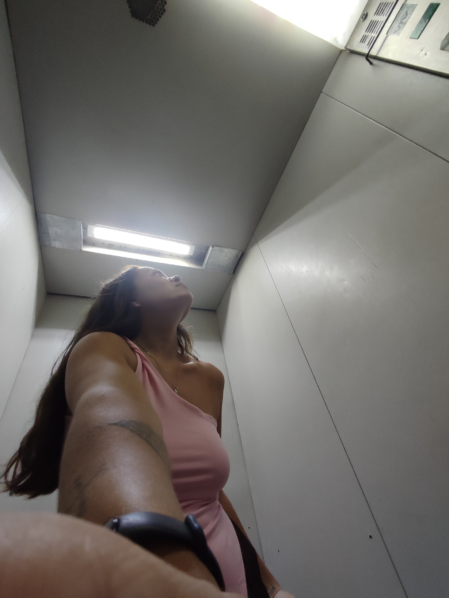 The image, taken in an elevator in the city of Chaikovsky, Russia, shows a low-angle shot looking upwards. A woman with long dark hair is visible, wearing a pink one-shoulder dress. She is looking up towards the ceiling. The elevator interior appears to be white or light-colored, with a fluorescent light fixture in the ceiling. A portion of the elevator's control panel, with buttons and indicator lights, is visible in the upper right corner. The woman's arm, holding what appears to be a selfie stick or camera, is in the foreground, partially obscuring her body. A tattoo is visible on her upper arm. The walls of the elevator show some minor scuff marks.