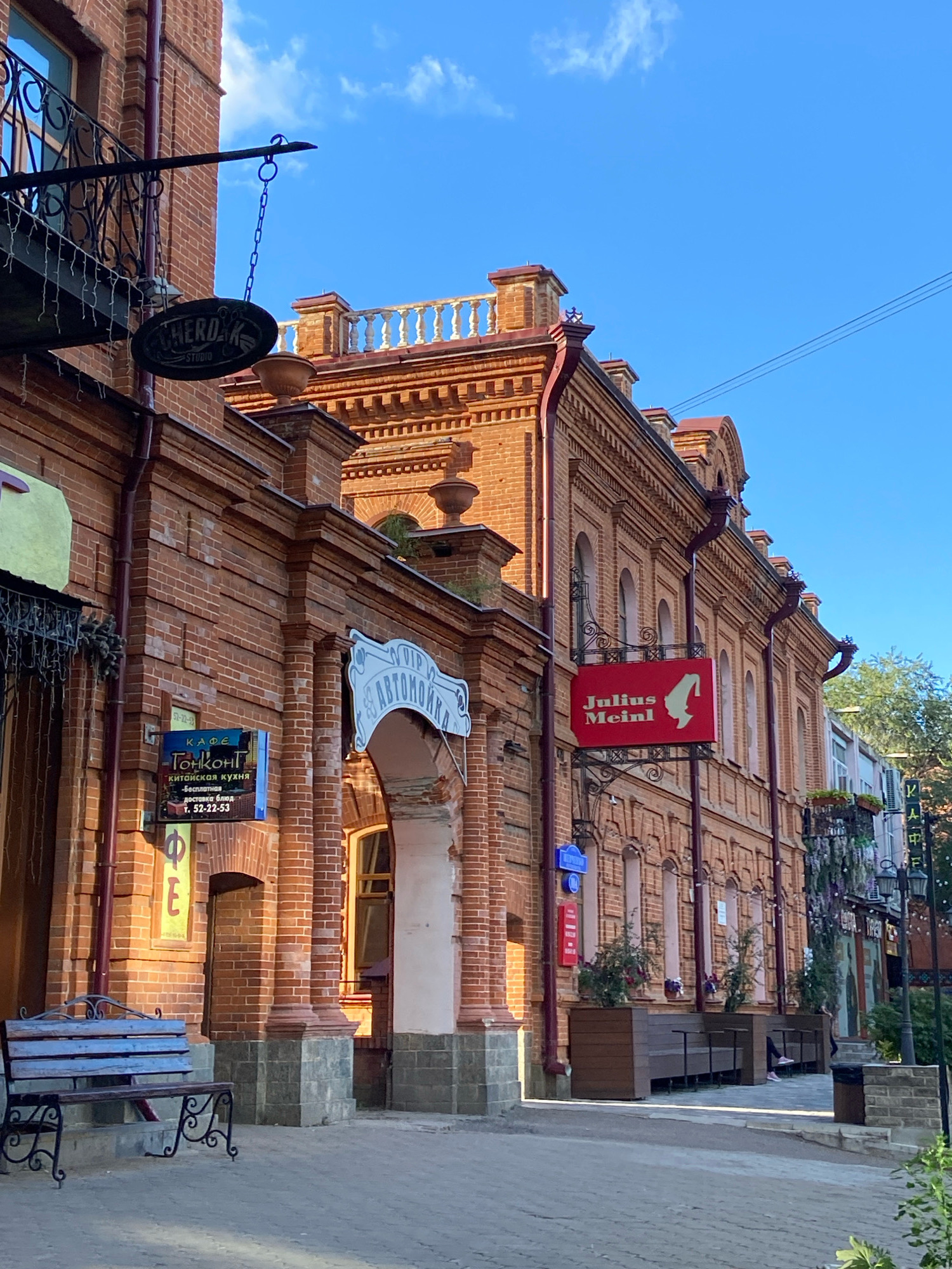 A low-angle, full shot captures a street view of historic brick buildings in Blagoveshchensk, Russia, bathed in the warm sunlight of a clear, blue sky. The architecture is characterized by ornate brickwork, arched windows, and decorative cornices, with a bright red sign for "Julius Meinl" prominently displayed on the right side of the frame. To the left, a dark oval sign reading "CHERDAK STUDIO" hangs from a wrought-iron bracket. Other signs visible include one for "КАФЕ Гонконг" (Cafe Gonkong), advertising Chinese cuisine and delivery, and a white arched sign for "АВТОМОЙКА" (Car Wash). A rustic wooden bench sits in the foreground, with planters containing greenery along the building's facade. The scene conveys a sense of urban charm and historical preservation.
