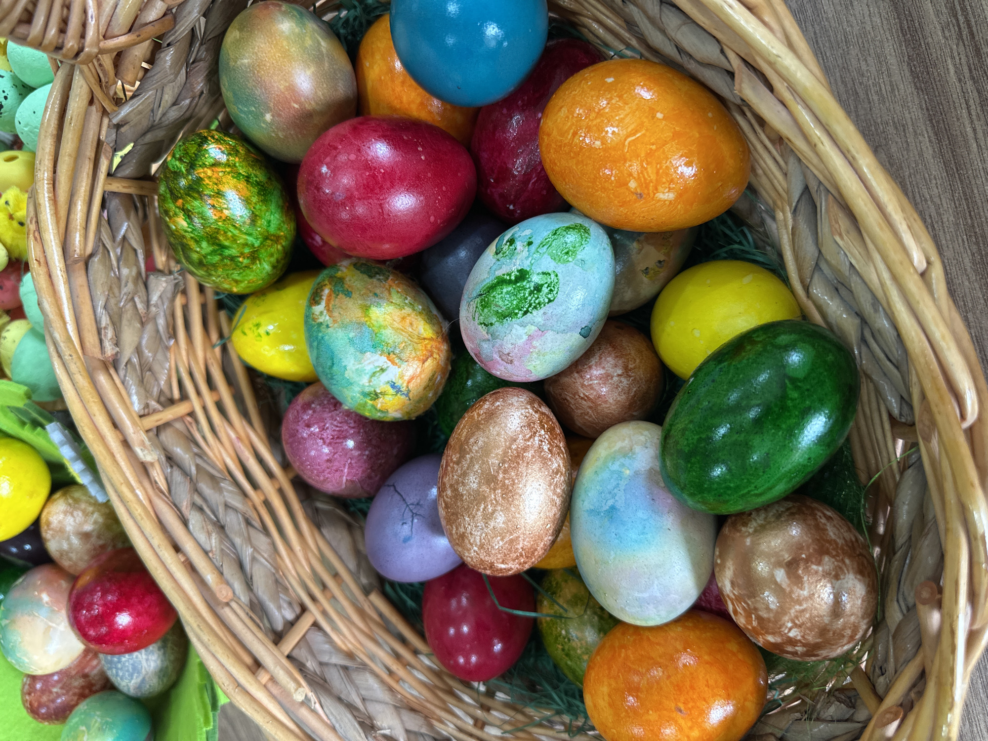 A wicker basket filled with approximately 25-30 Easter eggs of varying colors (red, orange, yellow, green, blue, purple, brown) and decorative styles is situated on a wooden surface. The eggs are nestled amongst strands of green grass-like material. The basket is made of light brown woven material. The surface beneath the basket is a light brown with a subtle wood grain pattern. The overall scene suggests an Easter celebration. The location is not discernible from the image.