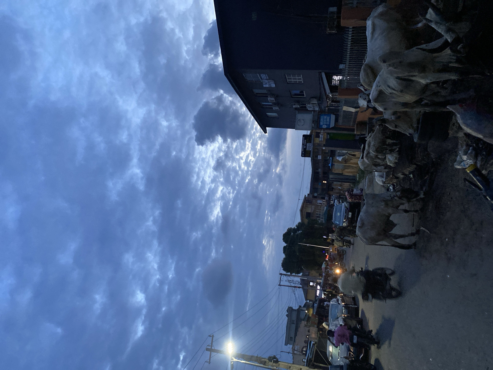 The image depicts a street scene in Lagos, Nigeria at dusk. The sky is filled with dense, dark clouds. A multi-story building dominates the left side of the frame, with signs and air conditioning units visible. The street is congested with vehicles, including cars, motorcycles, and what appear to be cattle being herded. Pedestrians are also present, some riding motorcycles. Power lines run along the street.