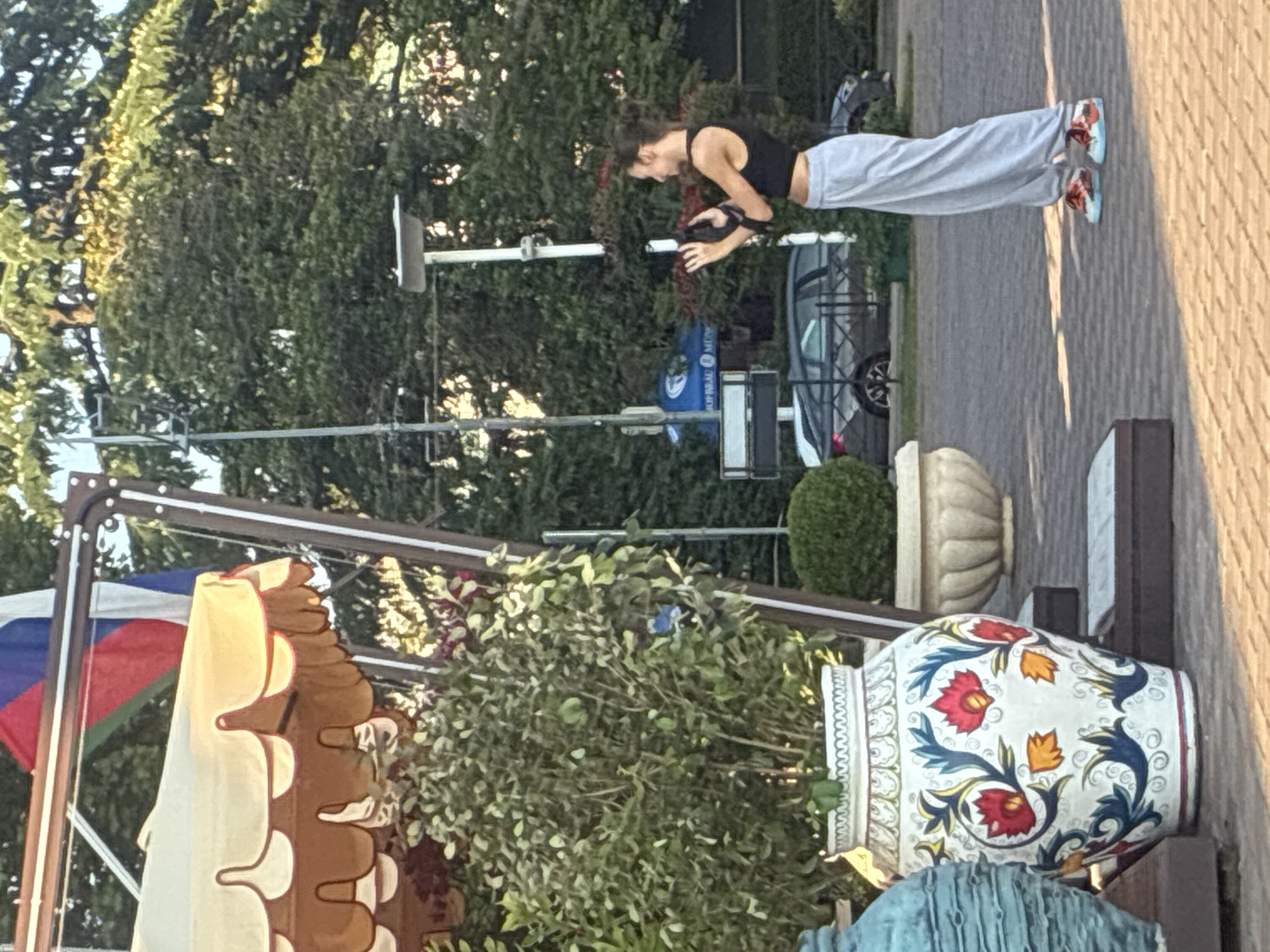 The image captures a vertical view of a young woman with dark hair, wearing a black tank top and light grey sweatpants, engaged in an activity outdoors. She is leaning forward, holding a dark object with straps, possibly a bag or camera. Her posture suggests concentration or interaction with the object. She is wearing colorful sneakers with orange and blue accents.
The setting appears to be a paved area, likely a walkway or plaza, with a stone pattern visible on the ground. Lush green trees form a dense backdrop, suggesting a park or garden area. Sunlight is casting strong shadows, indicating it is daytime, possibly late afternoon given the warm, golden hue of the light.
In the foreground, to the left, a colorful structure with a scalloped brown edge and a yellow canopy is partially visible. Flags, including a Russian flag, are also present near this structure. To the right, a large decorative ceramic vase with a white base and intricate floral patterns in red, blue, and yellow is prominently displayed. Another blue ceramic object is visible below the vase. A small, round green bush is also in the vicinity of the vase. In the background, a silver car is parked.
The overall scene seems relaxed and casual, with the focus on the woman and her activity, set against a backdrop of greenery and decorative elements. The image's orientation is vertical, with the subjects appearing to be standing on the right side of the frame. No text is clearly legible in the image.