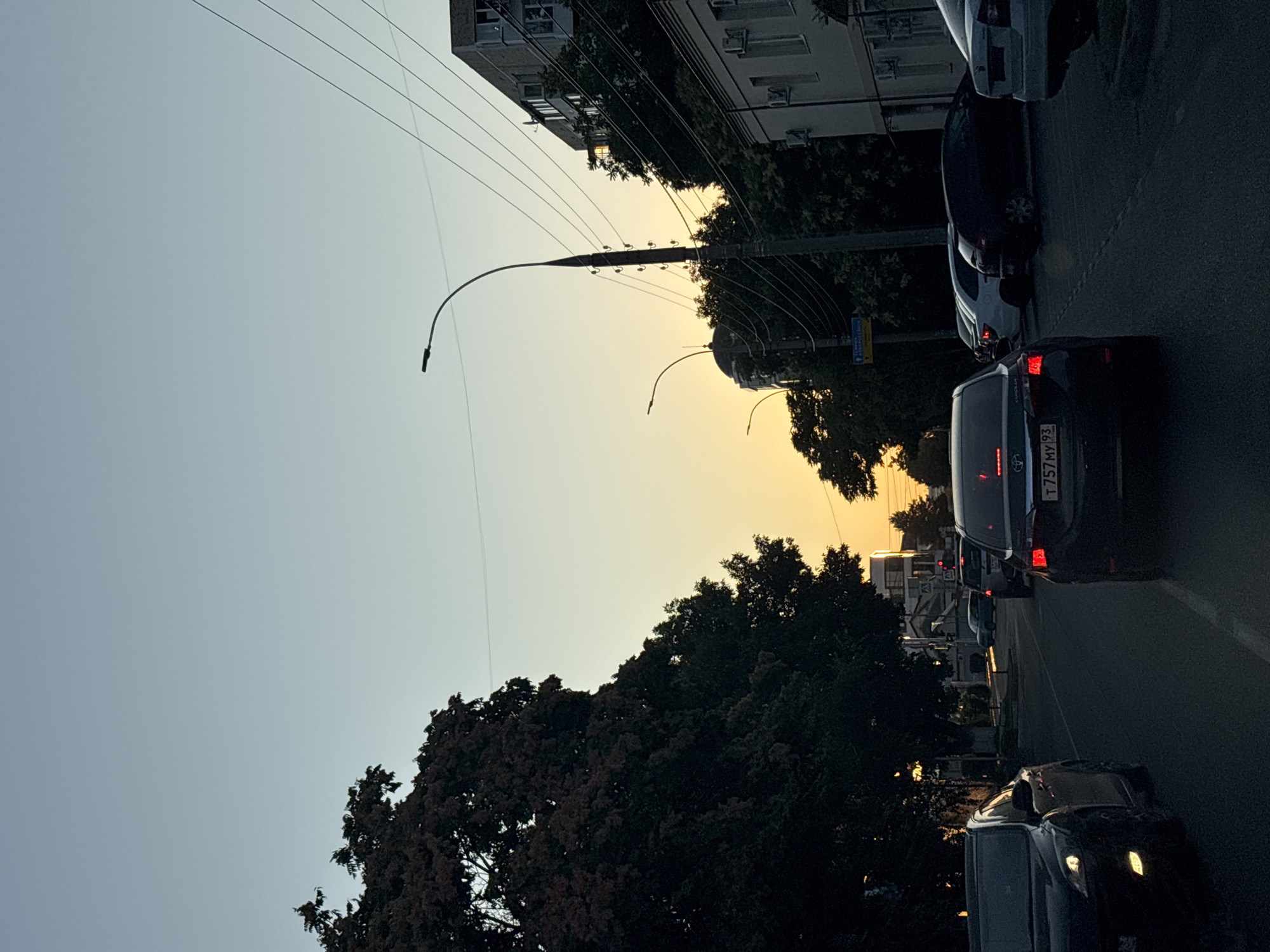 The image depicts a street scene at dusk in Krasnodar, Russia. The sky is a gradient of pale blue to warm yellow as the sun sets. Several cars are visible on the asphalt road, some in motion and others stopped, likely due to traffic or a red light. On the left, a dark sedan is in the foreground, its headlights illuminated. Further down the road, a black sedan with its taillights illuminated is in the center of the frame, displaying a license plate that reads "T 757 MY 93". Other vehicles, including a white sedan, are parked or driving further down the street.
Large trees with dark foliage line the left side of the road, casting shadows on the street. Buildings are present on the right side of the image, including a multi-story apartment building with windows and balconies, and another building with white facade and air conditioning units. Streetlights are visible, with one tall pole supporting multiple power lines. The environment suggests an urban setting during the evening hours.