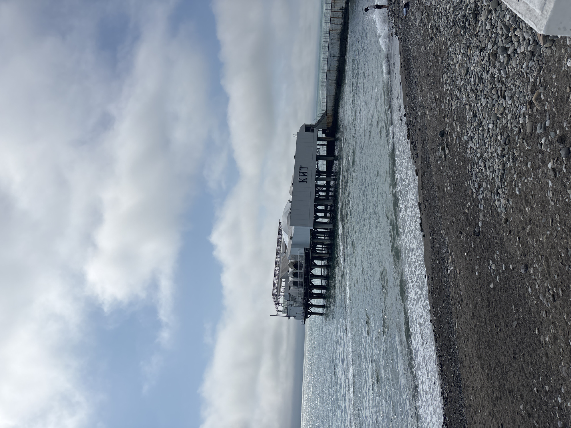 The image shows a pier in Sochi, Russia, with a building on it, adjacent to a pebble beach and the Black Sea. The building on the pier has the letters "КИТ" displayed on its side. A concrete retaining wall separates the beach from the city. The sky is overcast with clouds. The angle of the pier to the shore is perpendicular.
