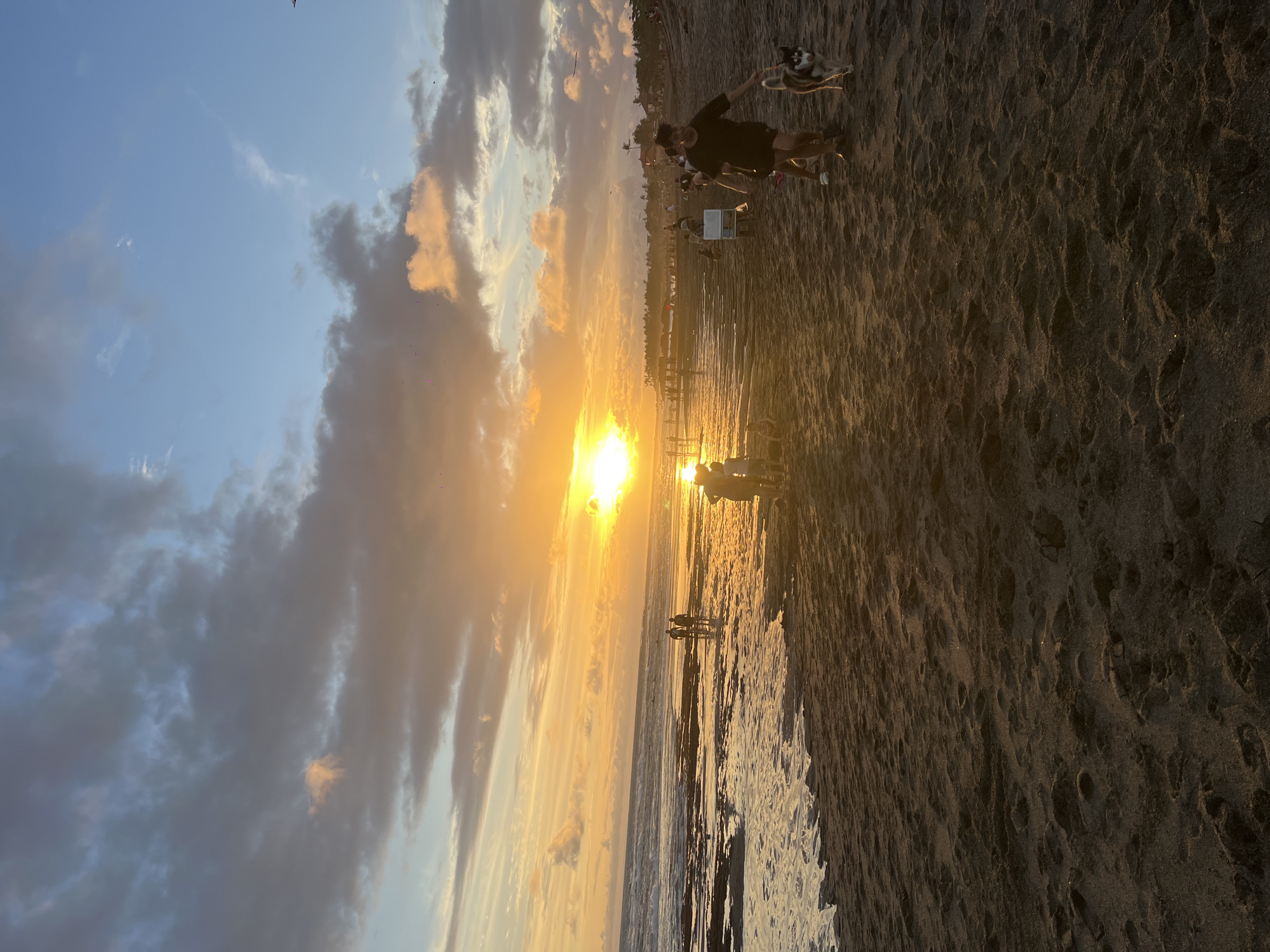 The image captures a sunset at a beach near Mengwi, Indonesia. The sun is setting near the ocean horizon, casting golden light across the sky, clouds, and beach. People are scattered along the shore, some walking near the water, others standing or sitting closer to the beach area. One person is actively interacting with a dog. The beach appears to have dark sand.