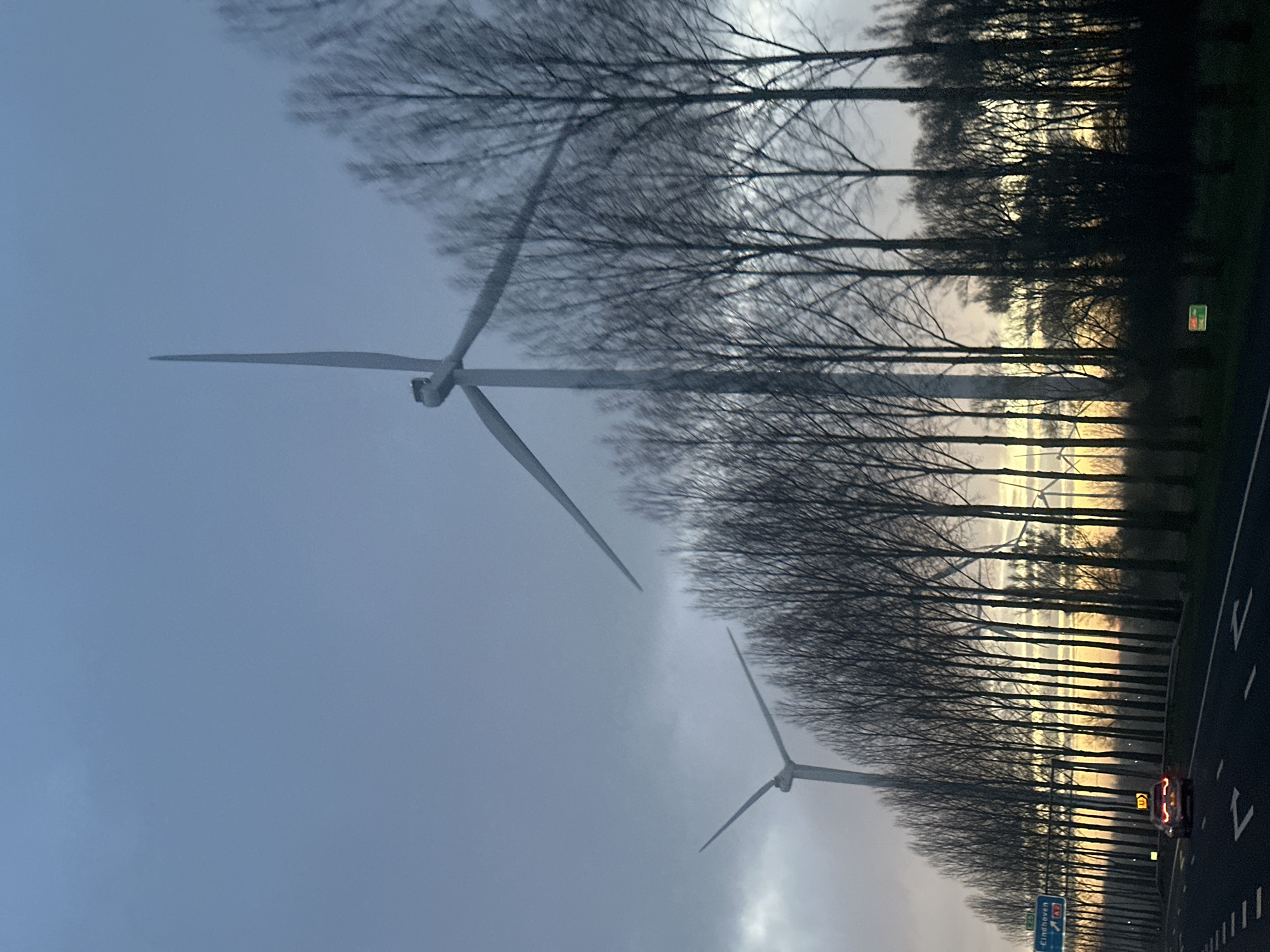 The image shows a highway scene in Enspijk, Netherlands. Two wind turbines are visible in the background, behind a line of bare trees. A car with illuminated taillights is traveling on the road. A road sign is visible in the lower right corner. The sky is overcast, with a hint of light on the horizon.