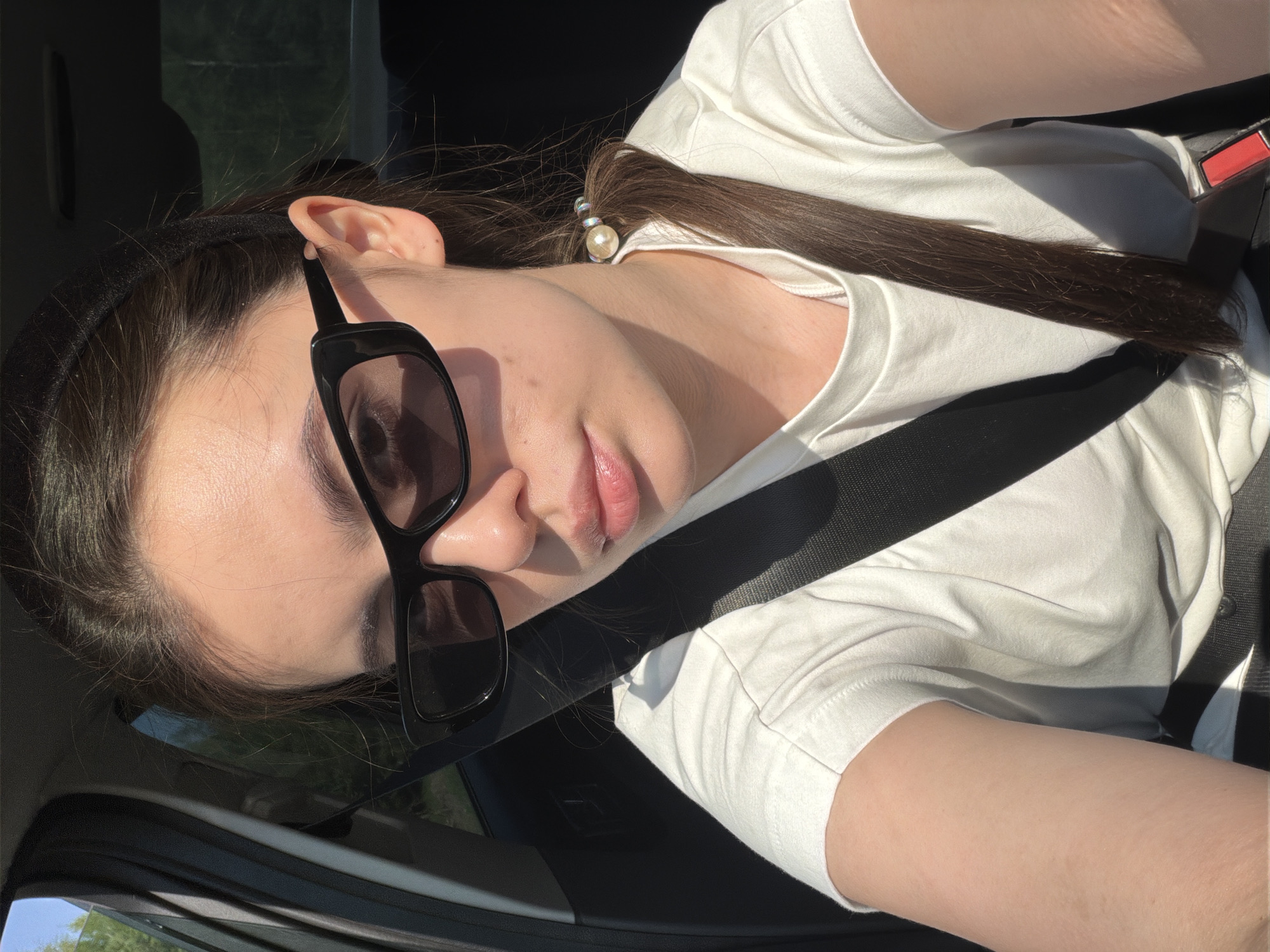 A woman with fair skin, wearing black-framed sunglasses and a white t-shirt, sits in a car, restrained by a black seatbelt. Her dark brown hair is pulled back, secured with a hair tie featuring a pearl-like embellishment. The interior of the car is visible, along with a glimpse of the outside through the window. The location is likely a vehicle within the city of Новая Казмаска, Russia.