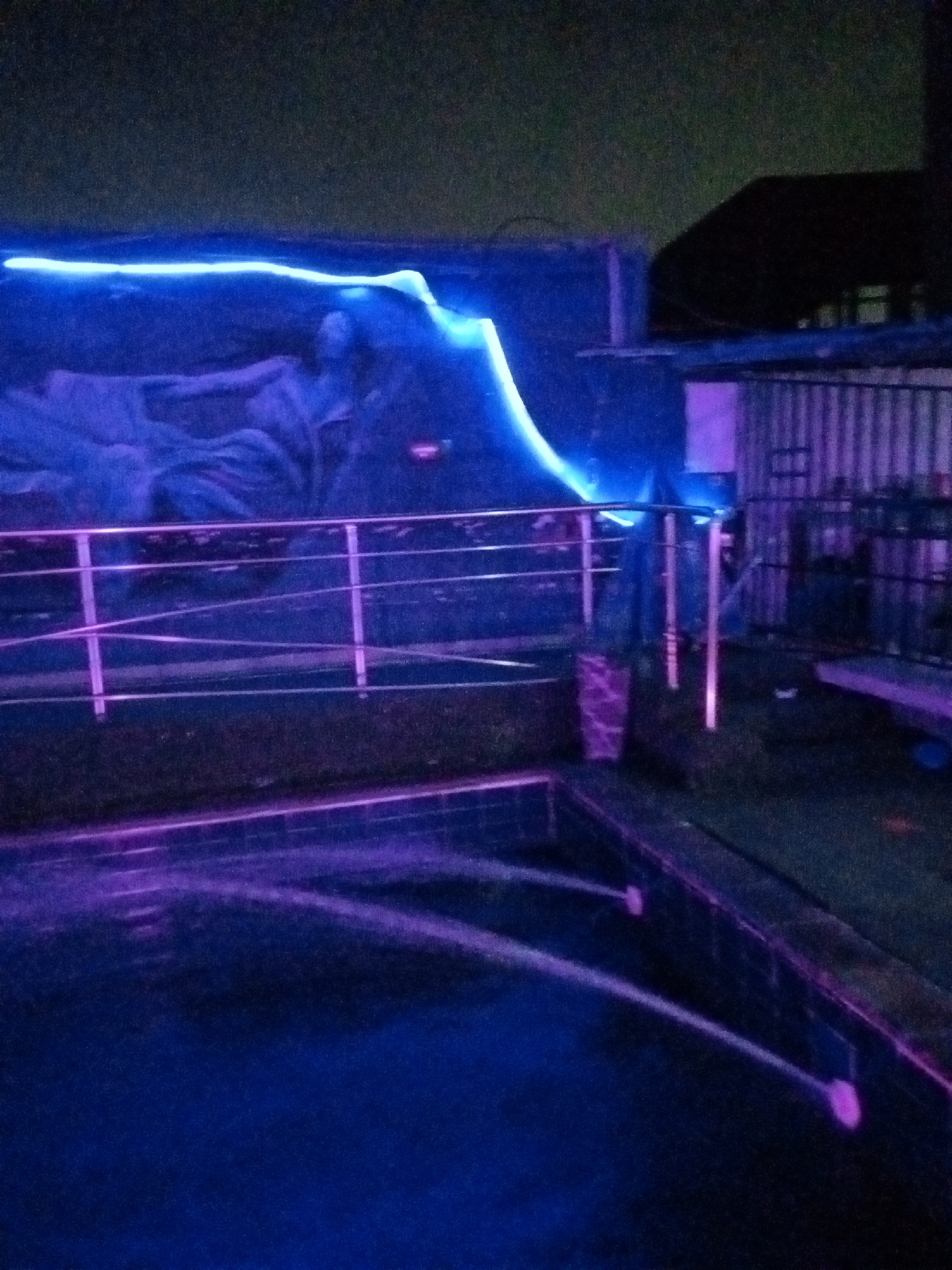 The image depicts an outdoor night scene, likely a poolside area, illuminated by artificial lighting. A swimming pool with dark blue water occupies the foreground, with visible jets creating ripples and spray. The pool area is bordered by a grassy lawn. A metallic railing runs along the edge of the lawn, separating it from a paved or tiled area. Behind the railing, a wall is visible, adorned with what appears to be a mural or a painted backdrop, enhanced by bright blue neon lighting that traces a wavy pattern across it. The background shows structures, including a building with a corrugated metal facade and a dark, silhouetted roof. The overall ambiance is moody and atmospheric due to the colored lighting, suggesting a recreational or entertainment venue in Ado, Nigeria. No people are clearly visible in the frame.