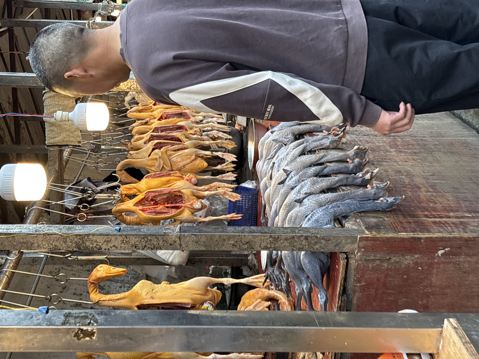 The image displays a bustling market stall in Jiangjin, China, featuring a vendor or customer amidst various prepared meats and aquatic products. A man, seen from a partial back and side view, is positioned towards the center-left. He wears a dark gray or brown long-sleeved shirt with a distinctive white and light gray diagonal stripe pattern on the side, and black pants. The text "RICKEY HAL" is visible on his shirt. His hair is short and appears to be dark with some graying.
The stall prominently showcases numerous whole, plucked ducks or poultry. Many of these yellow-skinned birds are split open, revealing their internal cavities and red meat, and are meticulously hung on metal skewers or hooks arranged on multi-tiered metal racks. One duck's head, complete with a yellow beak, is clearly visible among the hanging poultry.
To the right of the hanging ducks, a large pile of gray-skinned, elongated aquatic animals, resembling dried fish or eels, is neatly stacked on a wooden surface. These items are also prepared and displayed for sale. The entire scene is illuminated by several bright white LED light bulbs, suggesting an indoor market setting or a covered stall. The arrangement of goods on metal racks and wooden counters is typical of a bustling commercial market environment.
