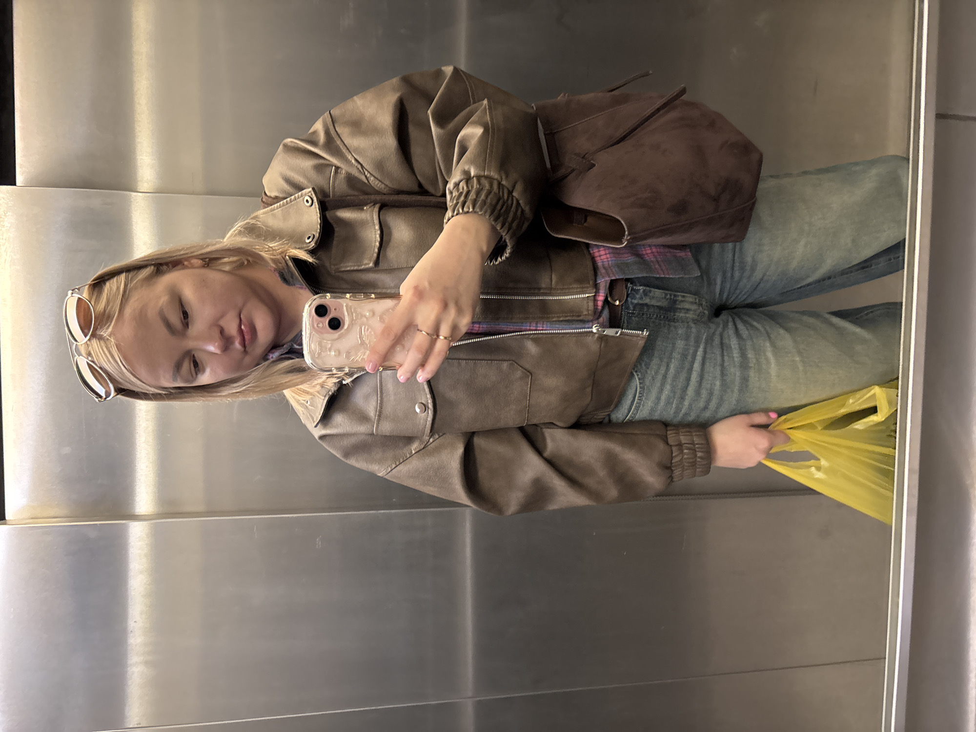 A woman is taking a selfie in an elevator with brushed metal walls. She has blond hair and is wearing sunglasses on her head, a brown leather jacket, a plaid shirt, and jeans. She is holding a cell phone in her left hand and a yellow plastic bag in her right hand. A brown suede bag is slung over her shoulder. The location is not discernable as Izhevsk, Russia, from the image.