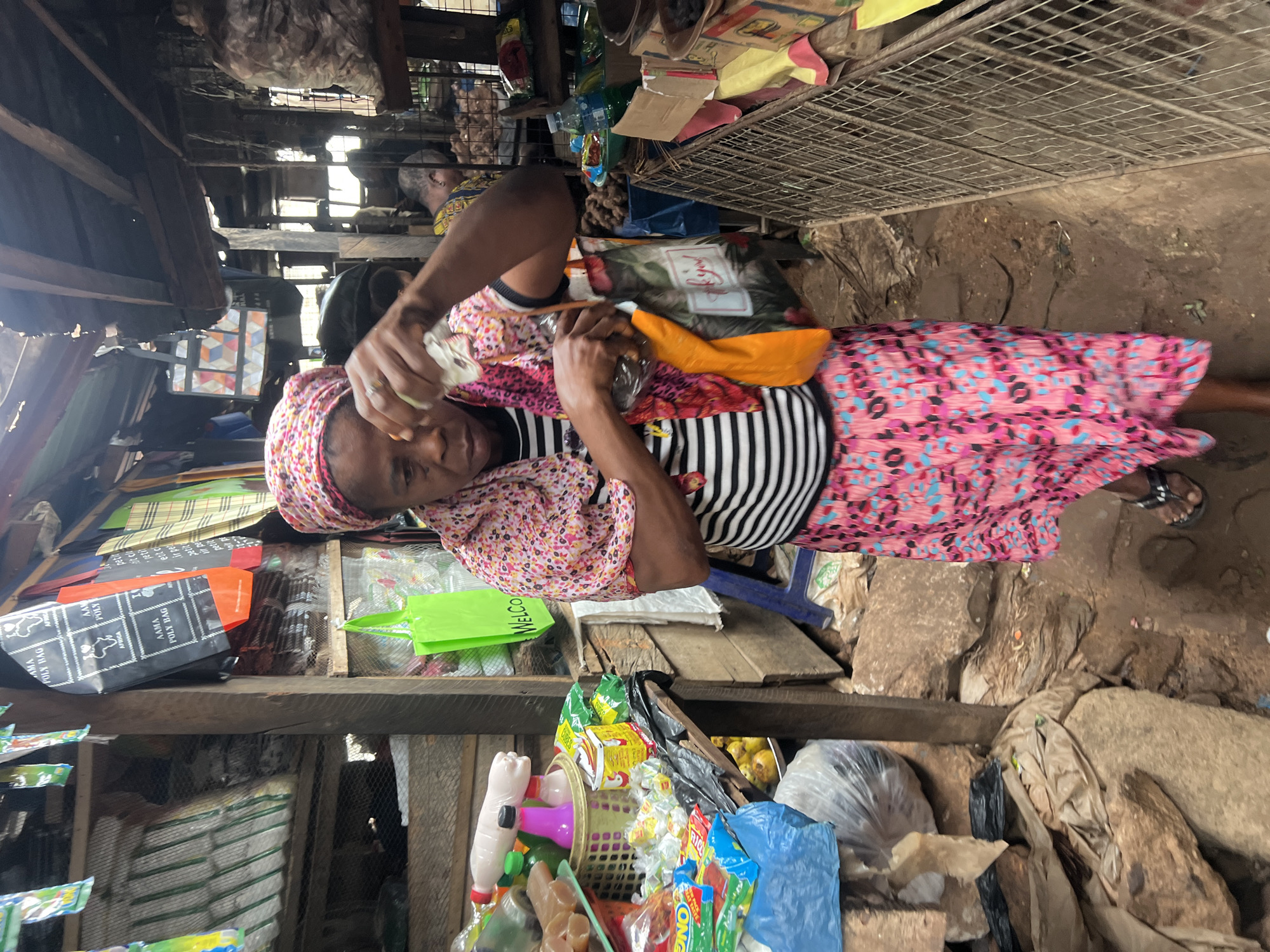 In Nkpor, Nigeria, a woman with a pink and white headscarf and a pink, blue, and black patterned skirt stands in a market stall. She wears a black and white striped shirt and black sandals. Her right hand, adorned with a ring, holds a clear plastic bottle, while her left hand grips an orange reusable bag patterned with floral motifs and the words "Thank You." The stall is filled with various goods, including plastic bags hanging from the ceiling, a basket of bottles and packaged candies, and bags of produce. The ground is uneven and covered in dirt and rocks. In the background, another person is visible behind the counter, partially obscured by the foreground action. The market appears to be constructed of wood and corrugated metal, with a caged area to the right containing more goods.