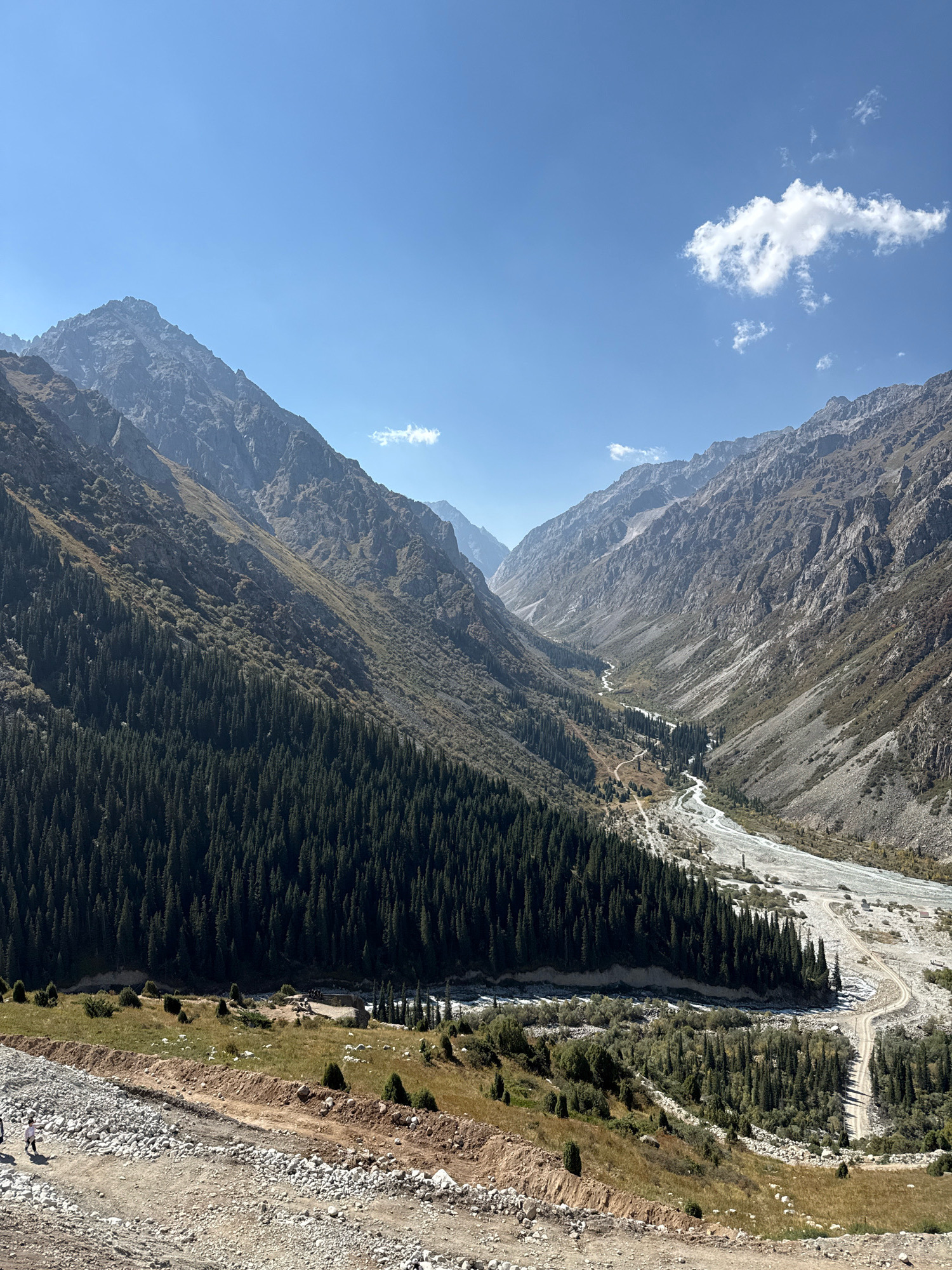 A wide-angle, high-angle shot captures a vast mountain valley under a clear blue sky with sparse white clouds. The valley floor is characterized by a winding, rocky riverbed and a dirt road. Dense evergreen trees cover the lower slopes of the mountains, transitioning to sparser vegetation and rocky terrain higher up. In the foreground, a dirt path with scattered rocks and sparse grass is visible, with two figures walking on it. The overall impression is one of a remote, natural landscape. The location is identified as the Ala Archa National Park, near Bishkek, Kyrgyzstan.