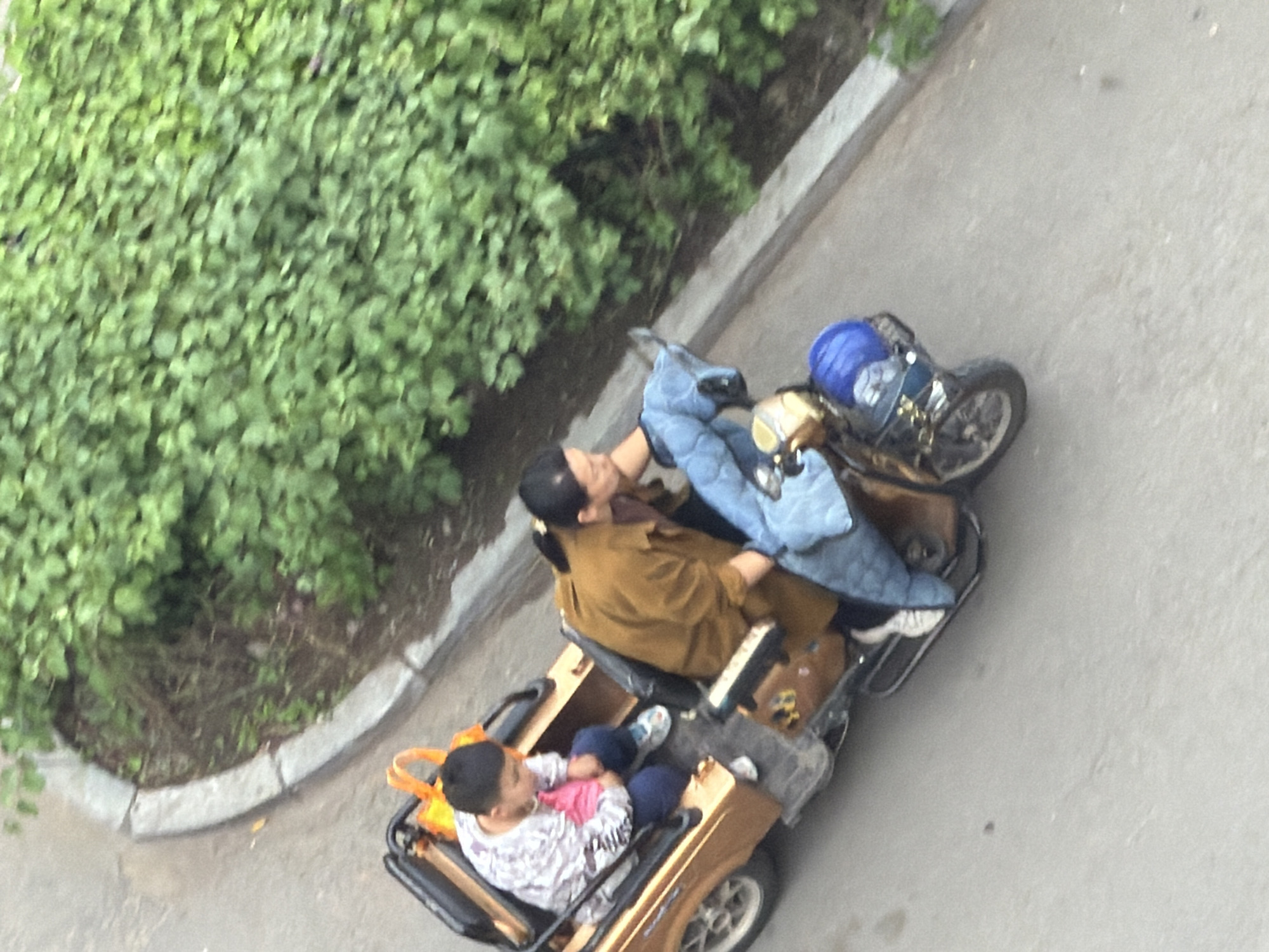 An overhead shot captures two individuals in a small, stationary electric vehicle on a paved surface next to a patch of green foliage and a concrete curb in Tongjiang, China. An adult, possibly a woman, is seated in the rear, wearing a brown jacket and dark pants, reclining with her head resting back. A child, likely a boy, occupies the front seat, dressed in a white patterned long-sleeve shirt and dark pants, holding a pink object. An orange bag is visible near the child's seat. Parked closely behind the electric vehicle is a dark-colored two-wheeled vehicle, possibly a scooter, with a blue cover or bag on its rear. The scene is illuminated by natural daylight, suggesting either overcast conditions or early morning/late afternoon, with no strong shadows. The individuals appear to be resting or paused on their journey. No discernible text is visible on any objects.