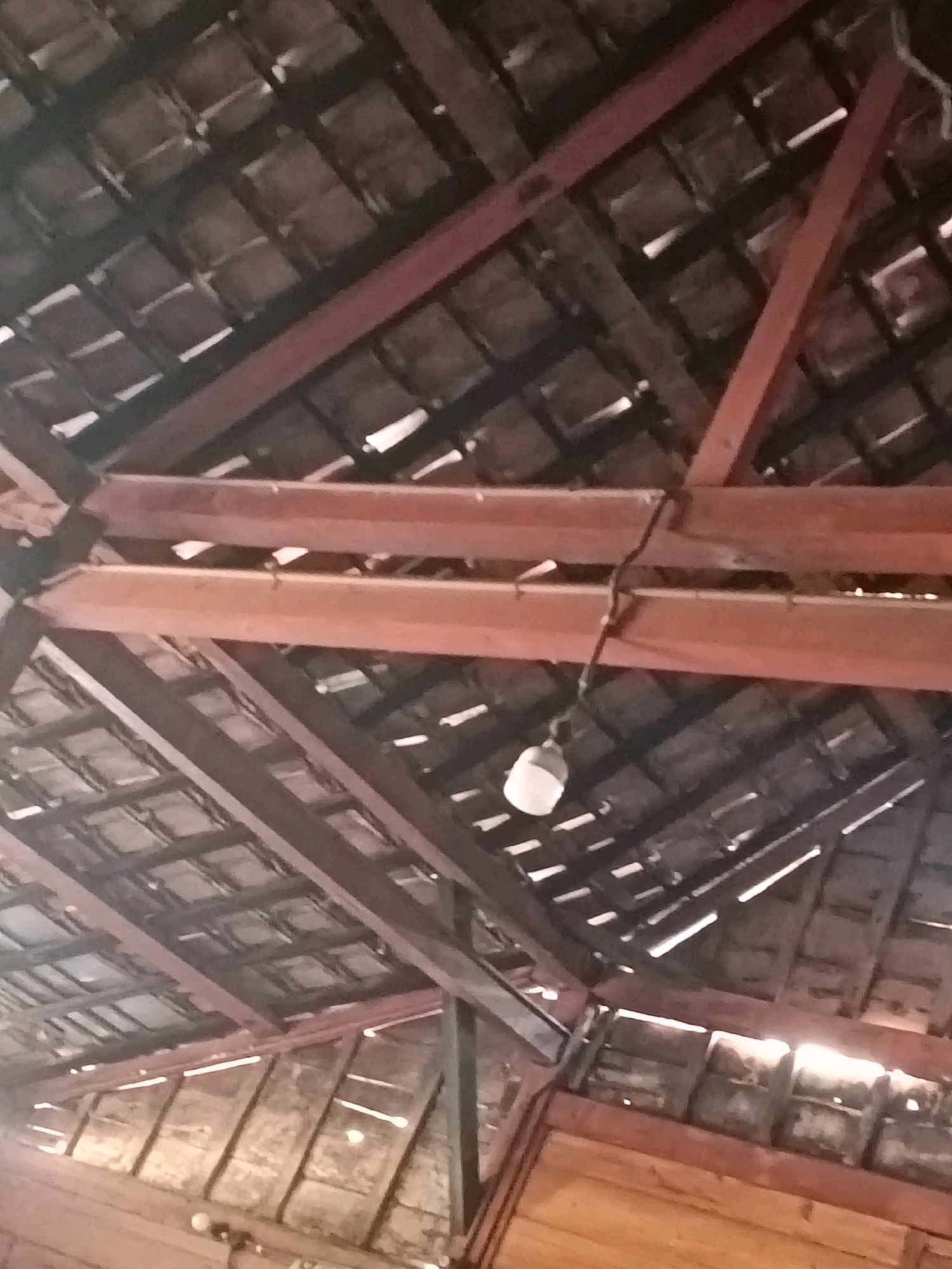 The image captures an upward view of a wooden roof structure, possibly part of a building in the city of Galuk, Indonesia. The roof is composed of overlapping, dark-colored tiles supported by a network of reddish-brown wooden beams and rafters. Gaps between the tiles allow natural light to filter through, creating bright lines. A single, bare light bulb is suspended from a wire attached to one of the beams. The lower portion of the image shows more wooden planks forming what appears to be the interior wall or ceiling of the structure. The overall impression is one of a rustic or traditional building.