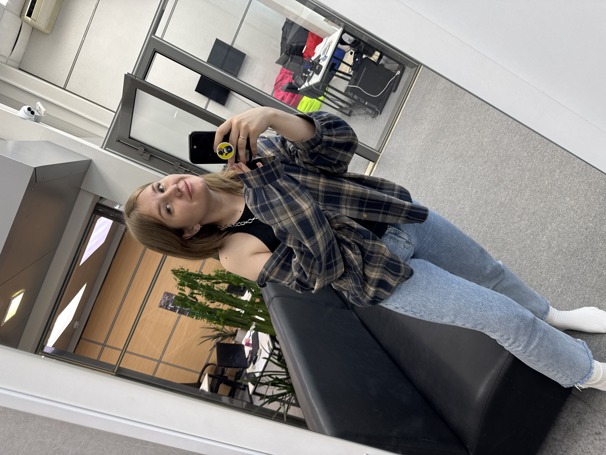 A woman is taking a selfie in a mirror. She is wearing a plaid shirt, jeans, a black top, white socks, and a silver chain. She is sitting on a black leather couch. The background shows an office space with windows, doors, a plant, chairs, desks, and a surveillance camera. Based on the location metadata, the image was taken in Perm, Russia.