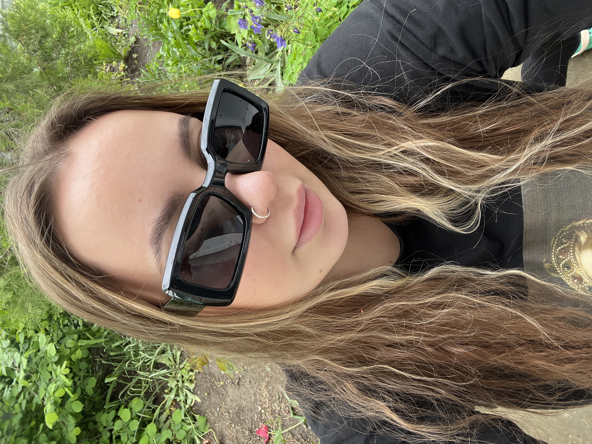 A woman with long, highlighted hair wearing black sunglasses and a nose ring lies in a grassy area. Her black shirt is visible. The location is not discernible from the image.