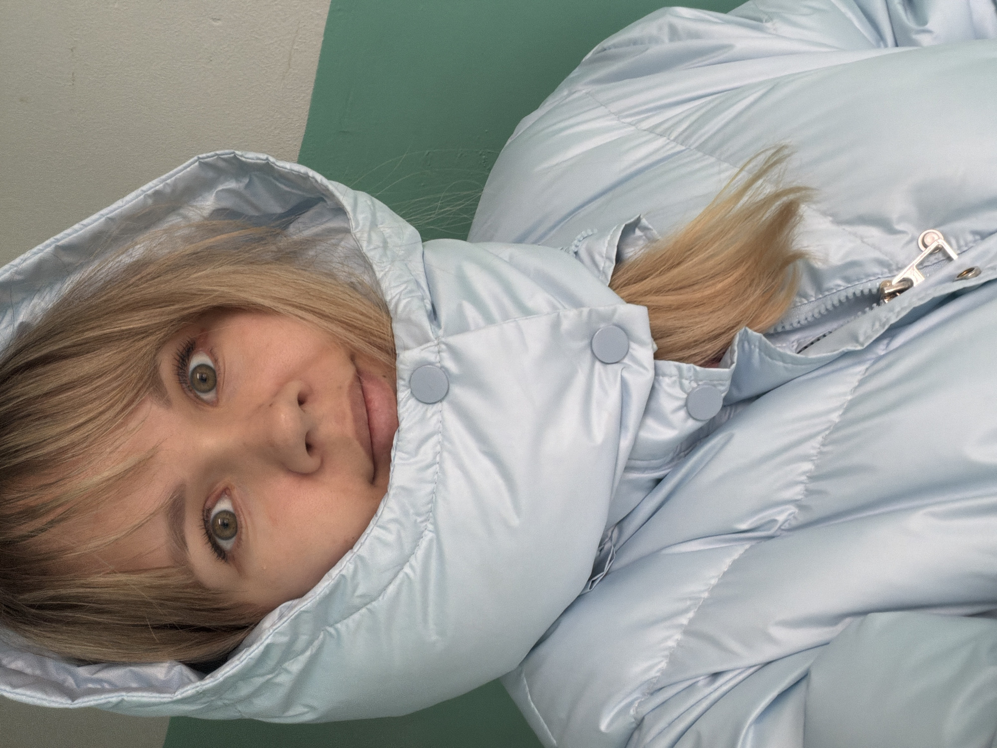 A young woman with blonde hair is posing for a picture while wearing a light blue puffer jacket with the hood up. She has fair skin, green eyes, and a playful expression with her tongue slightly sticking out. The background consists of a wall with two different colors, a lighter beige and a darker green, indicating an indoor setting. The location and city are unknown.