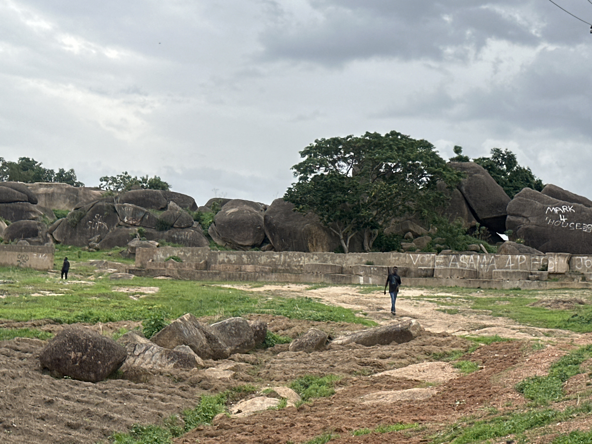 The image shows a landscape in Jos, Nigeria, characterized by large rock formations and sparse vegetation under an overcast sky. In the foreground, there is a rough, rocky terrain with patches of green grass and exposed soil. Scattered rocks of various sizes are visible.
In the midground, a low, concrete wall with graffiti divides the scene. One man is walking on a path in front of the wall, away from the viewer. Another person, standing alone, is located in the distant background.
Behind the wall are large boulders and trees, creating a natural barrier. Graffiti can be seen on the large rocks. The sky is gray and cloudy, suggesting an overcast day.