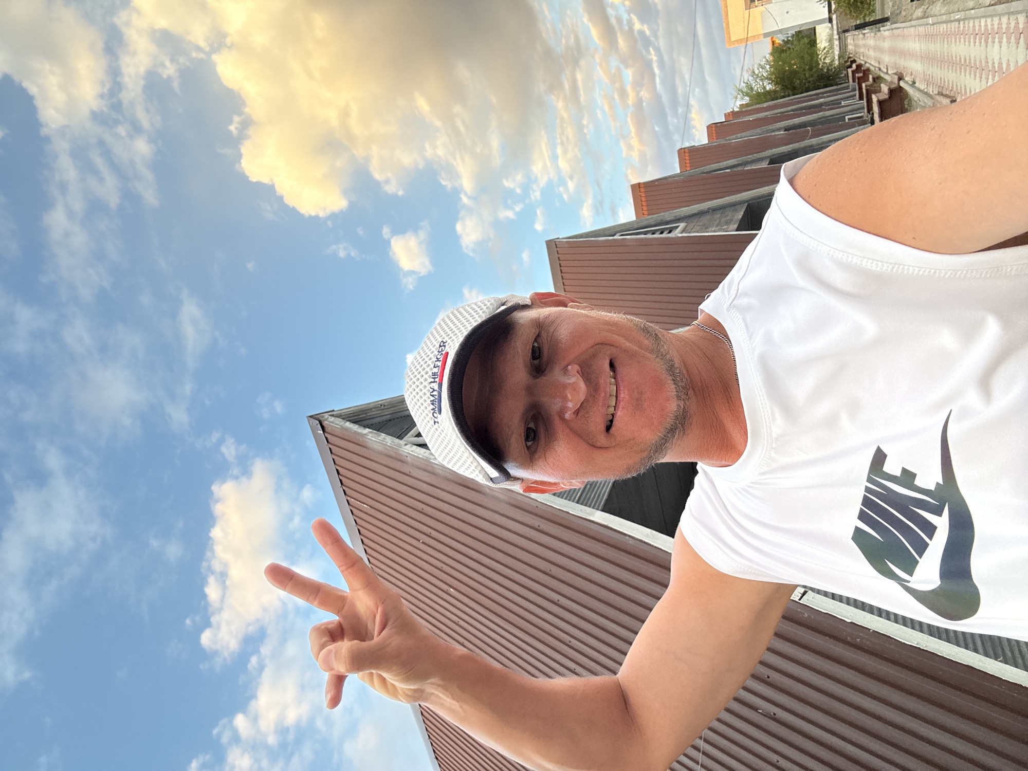A mid-shot, slightly angled selfie shows a man with fair skin, short brown hair, and stubble, wearing a white Nike sleeveless shirt and a white Tommy Hilfiger baseball cap. He is smiling and holding up his right hand with two fingers extended in a peace sign. The background features a row of identical A-frame buildings with brown corrugated metal roofs, a paved walkway, and a cloudy sky. The image appears to have been taken in Zharbulak, Kazakhstan.