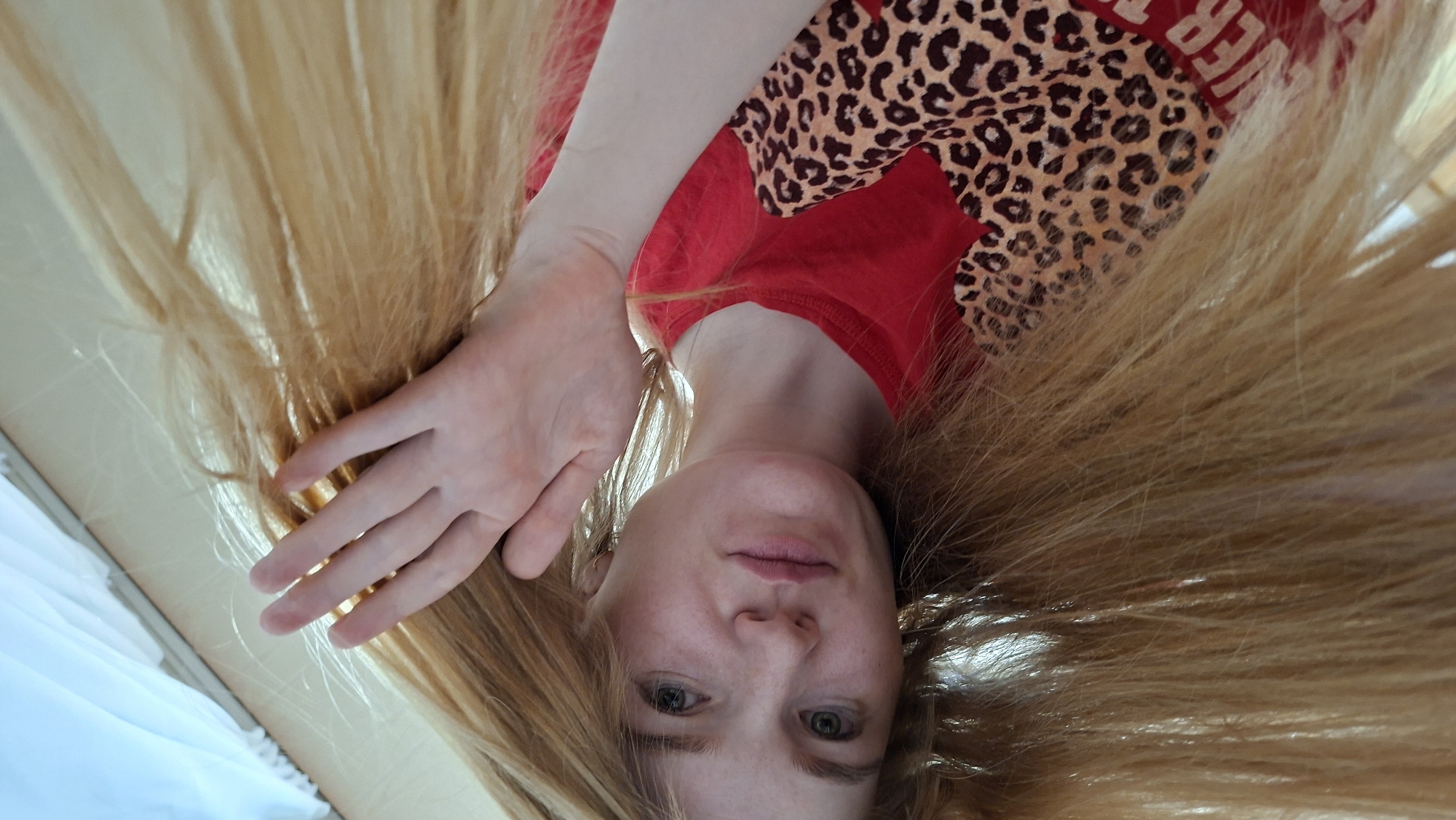 A young woman with long, blonde hair and fair skin with freckles is captured in a close-up shot. She is wearing a red t-shirt with a leopard print design on the chest area. Her right hand is raised, with fingers spread, touching her hair. The background appears to be an indoor setting with natural light filtering in, possibly from a window with white curtains. The image does not provide any specific visual cues that would allow for the identification of a location within the city of Большой Букор, Russia.
