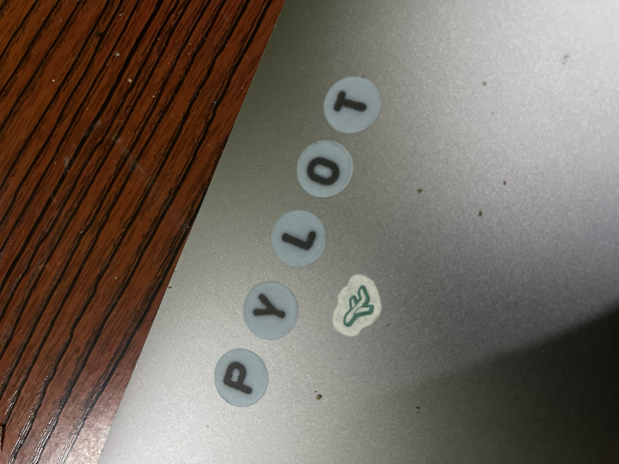 The image shows a close-up of a laptop surface with stickers. The stickers spell out "PYLOT" in individual round stickers. Next to the "Y" and "L" is a sticker of a stylized leaf. The laptop rests on a wooden surface. The location is not discernable. No people, actions, or interactions are visible.