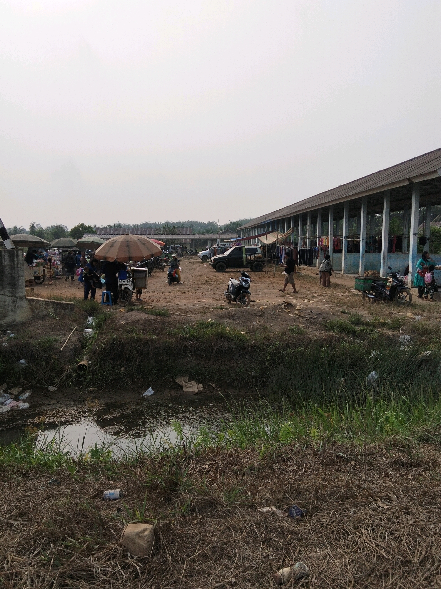 The image depicts an outdoor market scene in Perambahan Baru, Indonesia. In the foreground, a muddy ditch is filled with murky water and overgrown grass. Debris, including plastic bags, a can, and a fabric-wrapped object, is scattered on the dry ground and in the ditch.
The mid-ground shows a bustling market with several people, motorcycles, and market stalls. Two large, light-colored umbrellas are set up, providing shade for vendors and shoppers. A white motorcycle is parked under one of the umbrellas. People are seen walking, standing, and interacting with each other. One person appears to be holding a child. A few other motorcycles are visible, including one with a basket carrying produce. In the background, a long, open-sided building with a corrugated roof stretches across the scene, suggesting a more permanent market structure. Various vehicles, including a black pickup truck and a white car, are parked in the market area. Clothing is displayed for sale under the building's shelter. The ground is uneven and appears to be made of dirt and sand.
The background reveals more trees and buildings, and the sky is overcast, giving the scene a muted and hazy quality. The overall impression is of a lively, but somewhat unkempt, local market.