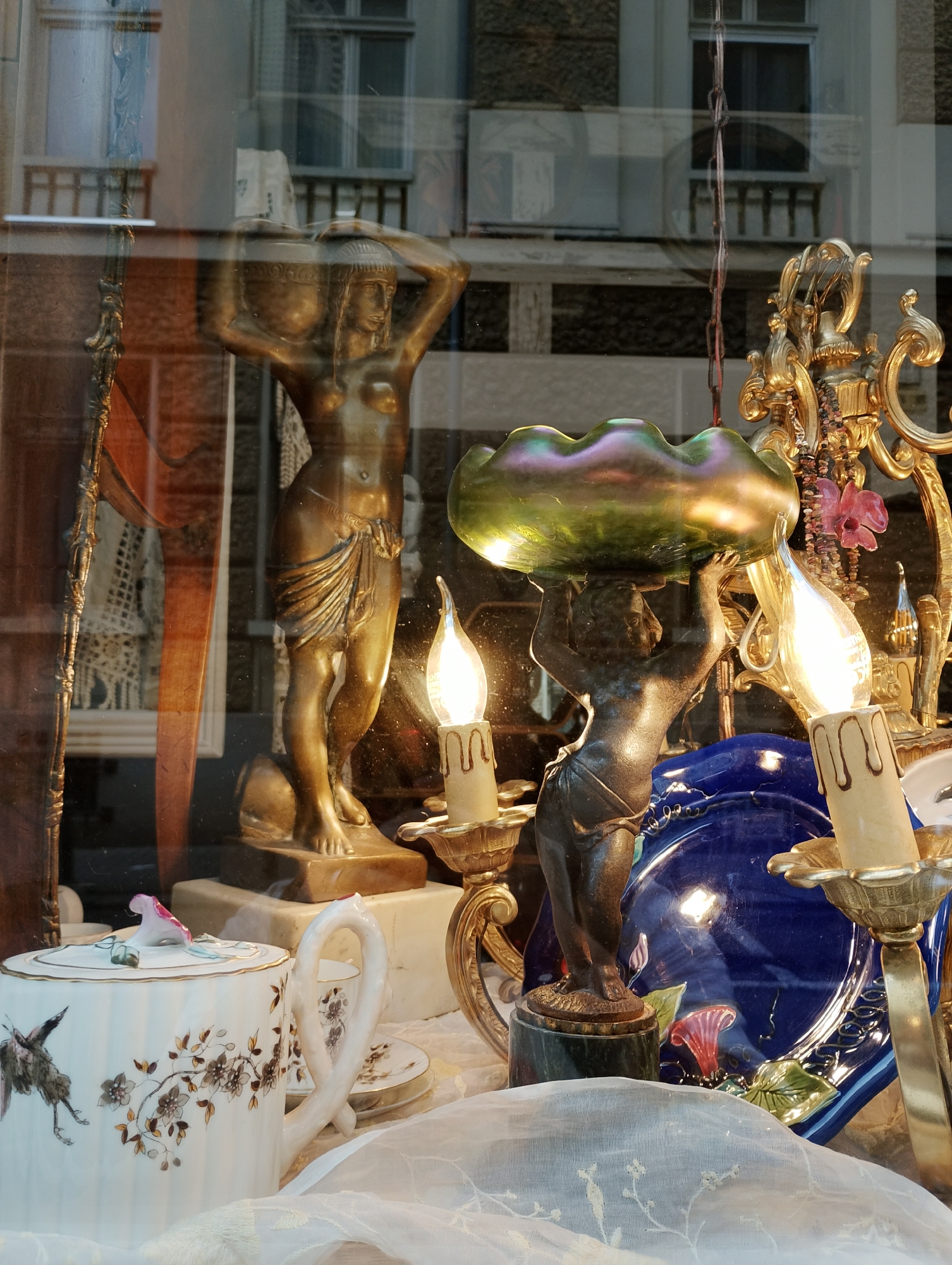 This media file captures a detailed view of a display window, likely belonging to an antique or specialty shop, showcasing an array of decorative objects. The scene is characterized by the prominent reflections on the glass, which superimpose elements of the exterior urban environment onto the interior display.
In the foreground, several ornate items are arranged. To the left, a tall, bronze-colored female statue, possibly an Atlantean or caryatid, stands gracefully, holding a large urn or basket on her head. Her lower body is draped in fabric. To her right and slightly lower, a darker, smaller statue resembling a putto or cherub holds an iridescent, wavy-edged bowl that shimmers with green and purple hues. Two electric light bulbs, shaped like flickering candles, illuminate parts of the display from golden, ornate holders. One of these holders is part of a larger, elaborate golden structure, possibly a candelabra, adorned with a hanging chain of beads and a small pink flower.
Further into the display, a vibrant blue ceramic plate with raised floral designs and a white porcelain teapot or sugar bowl featuring delicate brown and white floral patterns, along with a bird design resembling a crane or heron, are visible. The very bottom of the frame is softened by a sheer, light-colored draped fabric.
The reflections on the glass reveal multi-story buildings with windows and balconies, suggesting an urban setting during what appears to be daytime, though the interior lights are also on. There is no discernible text visible in the image. The overall impression is one of curated elegance and antique charm, with the reflections adding a complex layer to the composition.