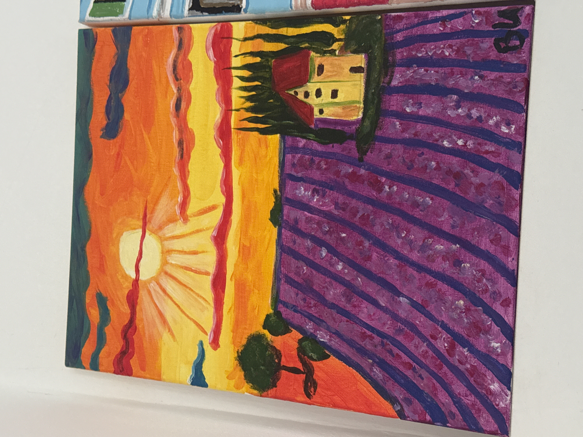 An eye-level, angled shot captures a square, vibrant painting hanging on a white wall. The painting is bisected vertically. The left half portrays a sunset scene, transitioning from green and blue at the edge to orange and yellow nearer the sun. The sun, painted in yellow, emits rays. Streaks of red and blue paint are visible. On the right side of the painting, there is a field of lavender depicted with purple and white strokes, with dark purple lines representing rows. A building, rendered in yellow with a red roof, sits atop a dark green area, possibly representing trees. In the lower central area, a small tree is painted.
There is no indication this painting is related to or located in Plovdiv, Bulgaria.