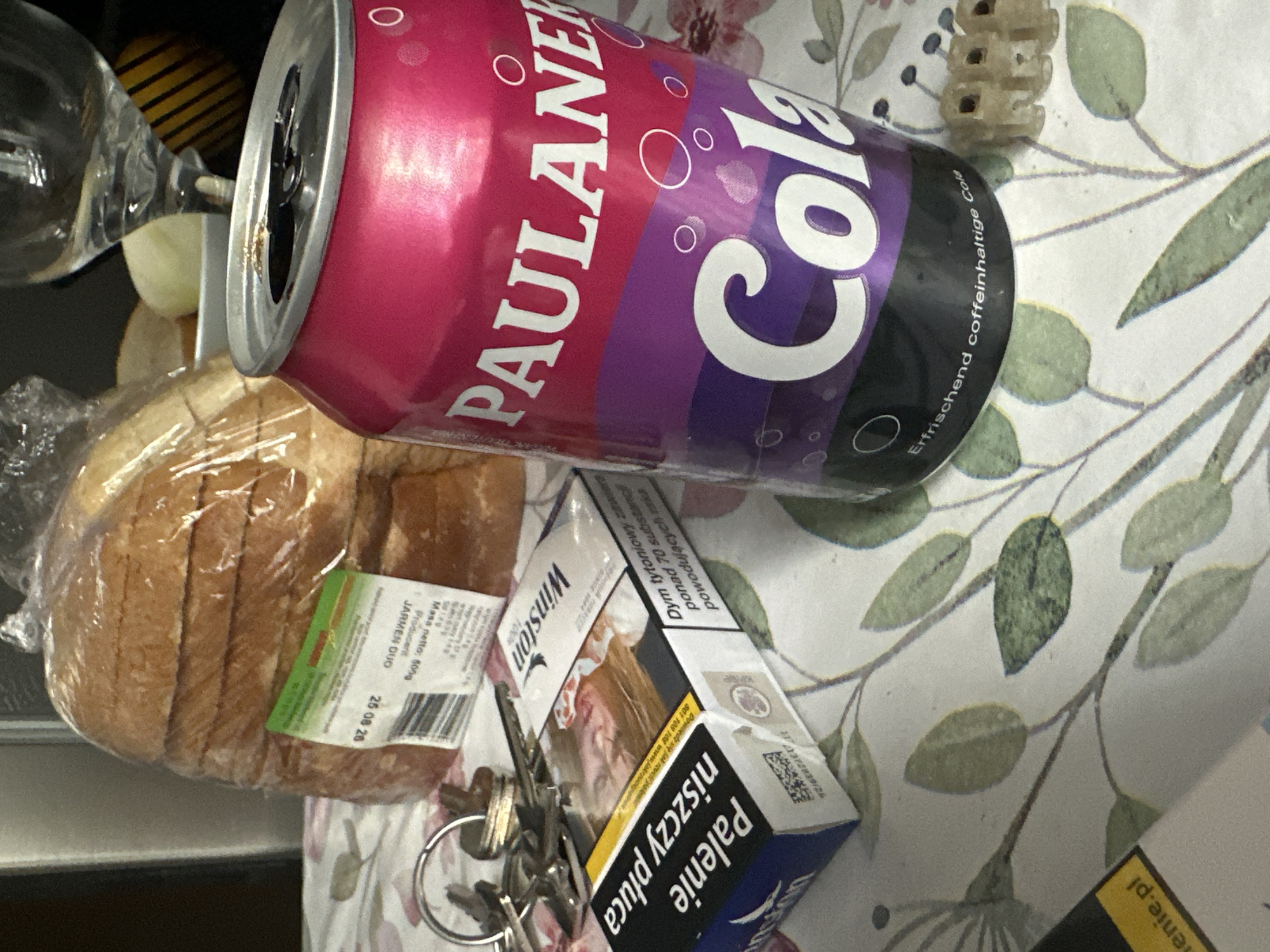 An indoor, eye-level, medium shot captures a collection of items arranged on a floral-patterned tablecloth. In the foreground, a can of Paulaner Cola is prominently displayed, its magenta and purple branding clearly visible. To the left, a pack of Winston cigarettes lies next to a set of keys. Behind these, a loaf of sliced bread is wrapped in plastic. A glass, likely for drinking, is visible in the background, partially obscured. The lighting suggests a domestic indoor setting. No specific location in Görlitz, Germany is indicated by the image itself.