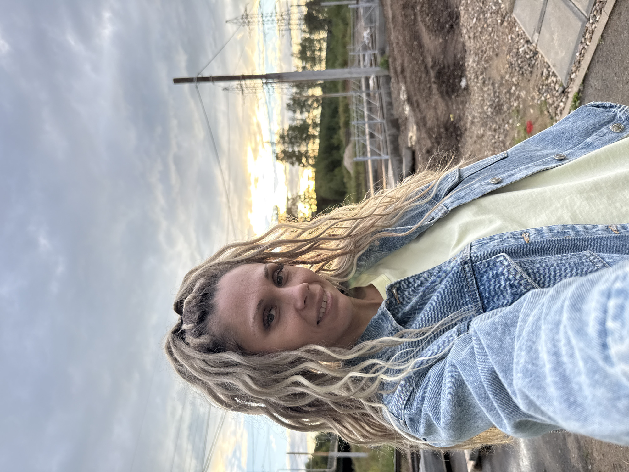 A woman with blonde dreadlocks is taking a selfie in an outdoor, possibly industrial, location. She is wearing a light yellow t-shirt and a blue denim jacket. The background shows a cloudy sky with hints of sunset light, trees, power lines, and construction debris. The location appears to be in or near Ignatovo, Russia, as suggested by the context.