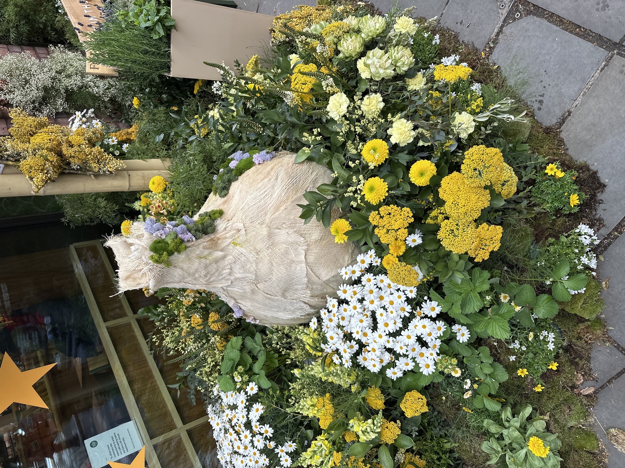 The image captures a floral arrangement in front of a building. The arrangement includes yellow, white, and green flowers, and some greenery. A light beige fabric structure is in the middle of the flower arrangement. The arrangement is set on what appears to be moss or grass, adjacent to a paved surface. Part of the glass front of a building is visible, with a notice in the window that mentions a café seating space, suggesting a restaurant or café setting. A building with decorations in the window is also visible in the background. The location is the city of London, United Kingdom.