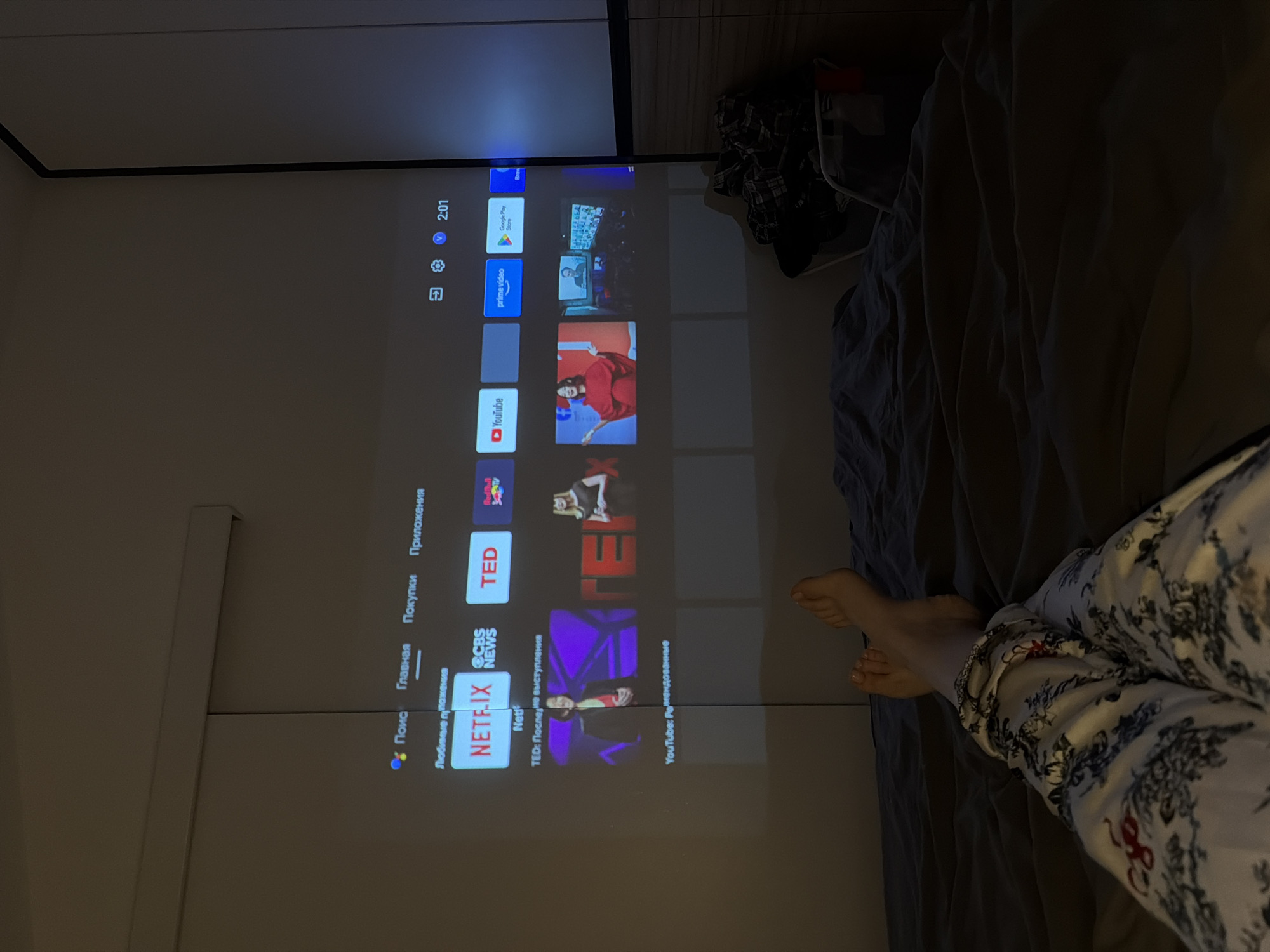 Here is a forensic description of the image:
**Scene:** A bedroom interior, viewed from an elevated position. A projection screen displays a smart TV interface on a wall. A person is relaxing in bed, visible from the knees down, wearing patterned pajama pants.
**Objects:**
* **Projection Screen:** Displaying a smart TV interface, featuring icons for Google Play Store, Prime Video, YouTube, Netflix, TED, and Red Bull. The interface language appears to be Russian.
* **Bed:** Covered with a dark gray/blue bedsheet.
* **Pajama Pants:** White with a dark floral print.
* **Shoes:** Black sneakers or boots.
* **Cupboard:** Light gray colored cupboard doors.
**People:**
* A person is visible from the knees down, wearing patterned pajama pants and is relaxing in bed.
**Environment:**
* The room appears to be dimly lit.
**Actions:**
* The person is relaxing in bed.
* The smart TV interface is displayed on the wall.
**Location:**
* City: Unknown
* Region: Unknown