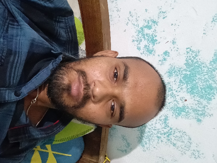 The image shows a man in a blue, striped shirt. He has a dark beard and mustache. His eyes are looking directly at the camera, with a serious expression. The man is sitting on a bed with a green and yellow patterned pillow behind him. The wall behind him is painted white with blue-green splotches. The photo was taken in an indoor setting in the city of Sambalpur, India, located at coordinates 21.468, 83.994. The image quality is good, and the scene is well-lit. The focus of the image is the man's face. The background is out of focus.