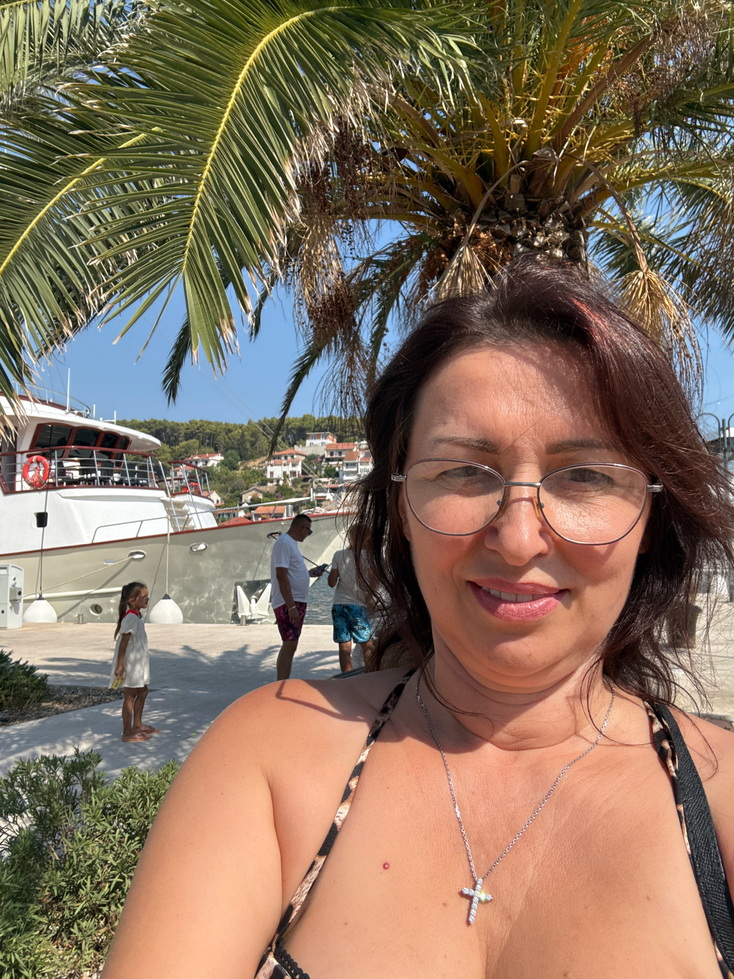 A woman takes a selfie on a sunny day in Jelsa, Croatia. She is wearing glasses and a leopard print bikini top. She is also wearing a silver necklace with a cross pendant. In the background, a large white boat is docked at a pier. A young girl in a white dress stands on the pier near the boat. Two men are also visible on the pier, one wearing a white t-shirt and patterned shorts, and the other wearing a t-shirt and blue shorts. Palm trees with fronds are in the foreground and overhanging the scene. Behind the pier, buildings with red tile roofs are nestled on a hillside, characteristic of Mediterranean coastal towns. The sky is clear and blue, indicating good weather. The overall scene suggests a vacation or a leisurely day by the water.