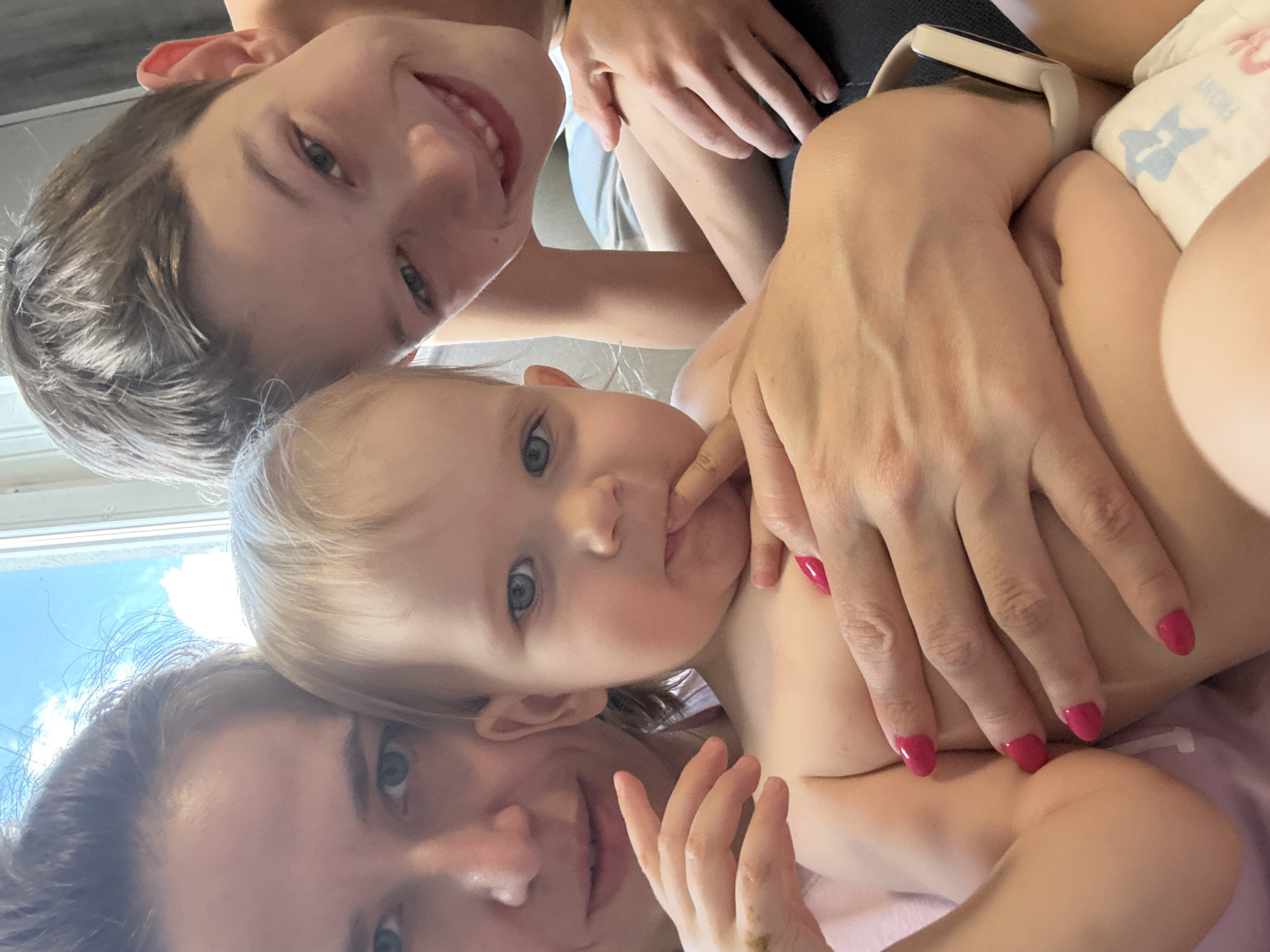 A close-up selfie taken from a high angle shows three people: a young child and two adults. The child, with wide blue eyes and blonde hair, is in the center, looking up with their finger to their mouth. To the left, an adult with brown hair and a wide smile is partially visible, with their head tilted. Below them, another adult's face is mostly seen, with their mouth open in a smile and their eyes looking directly at the camera. They have dark hair. The adults appear to be holding the child.
The scene is intimate and suggests a family moment. The lighting is bright, indicating it might be daytime. The background is blurred, but hints of a window with blue sky and clouds can be seen, possibly suggesting an indoor setting with a view of the outdoors. The overall mood is happy and affectionate. The adult on the right has bright pink nail polish on their fingernails. A white and blue diaper with a star pattern is visible on the child. A smartwatch with a light-colored band is also visible on the wrist of the adult on the right. No visible text is present in the image. The context provided places the location in Khimki, Russia.